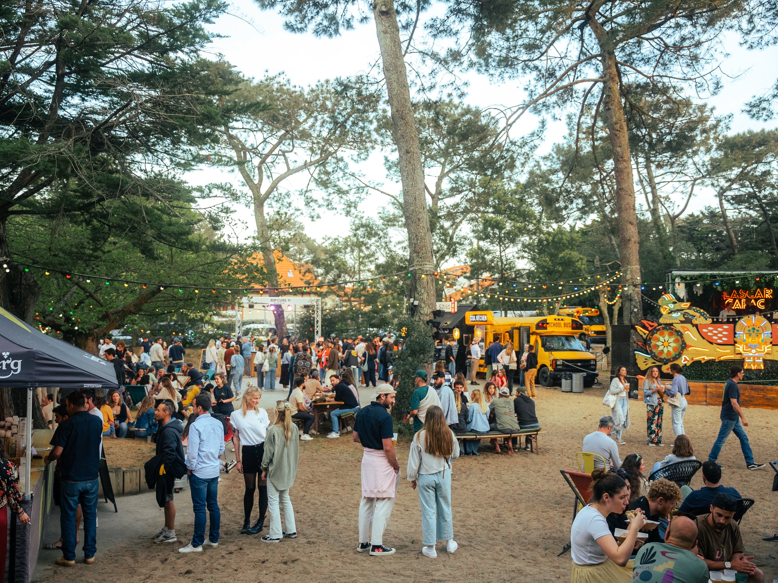 Une scène d'un parc ou d'une fête en plein air avec beaucoup de gens, des arbres, des guirlandes lumineuses et un stand coloré avec une carcasse de dragon.