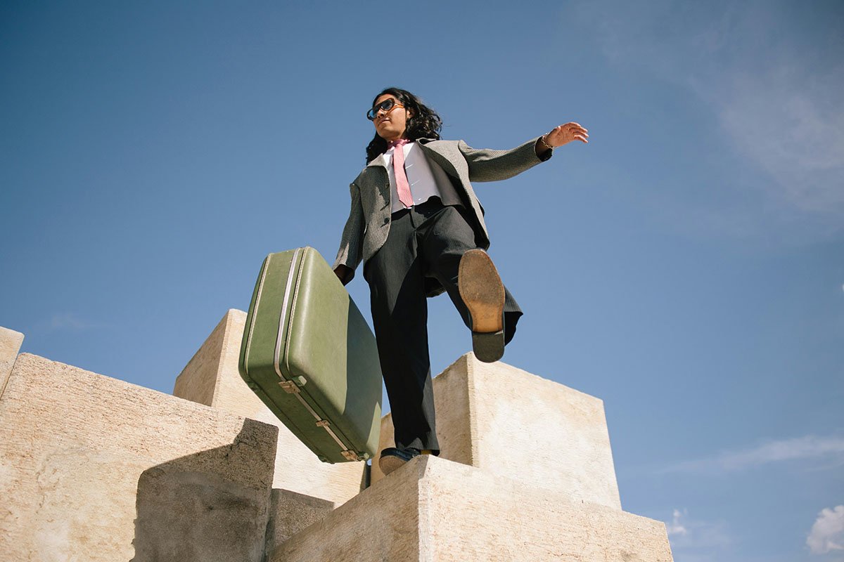 Femme en costume d'affaires avec valise verte, marchant sur des blocs de pierre sous un ciel bleu.