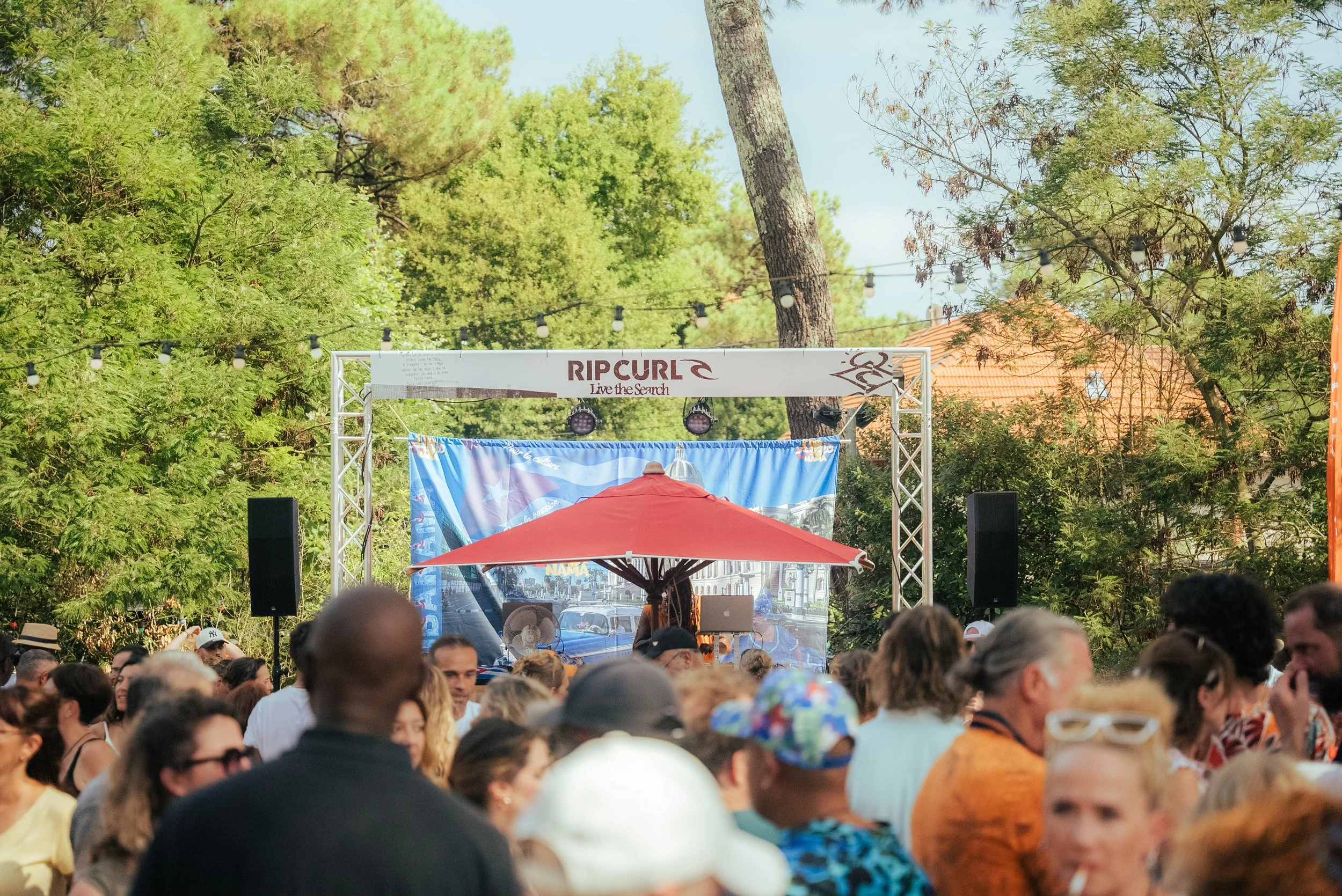 Un rassemblement de personnes lors d'un événement en plein air sous un ciel clair, avec un décor d'arbres et une scène équipée pour un spectacle ou un concert.