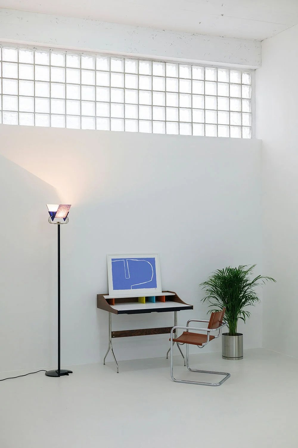 Intérieur minimaliste avec une lampe sur pied, une table design avec un tableau abstrait en bleu, une chaise en métal et un pot de plante verte devant un mur blanc lumineux par une grande fenêtre en blocs de verre.