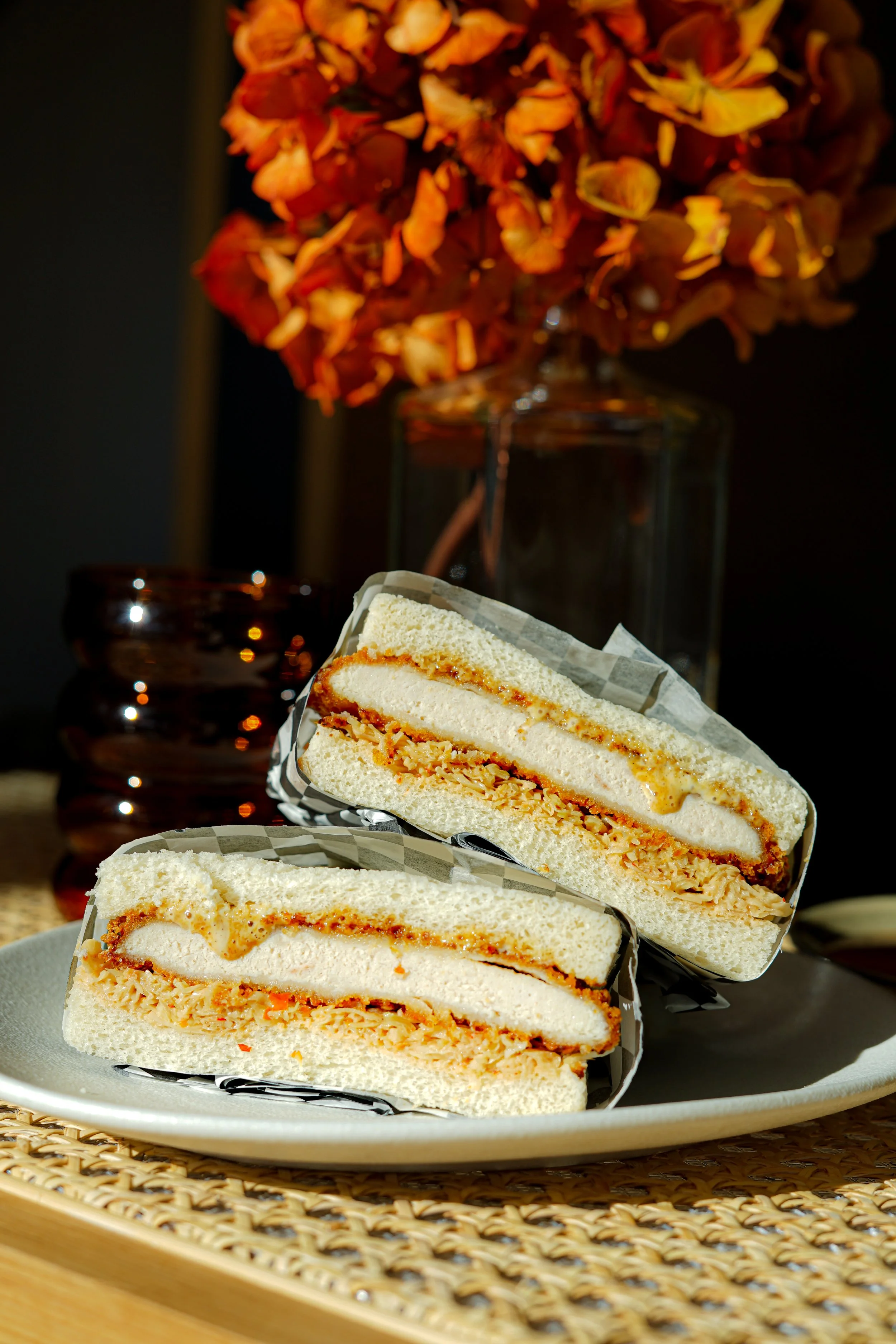 Gâteau en sandwich avec une couche de crème et une couche de pâte de noisette, présenté dans une assiette blanche. En arrière-plan, un bouquet de fleurs orange dans un vase en verre et un petit objet en verre foncé, sur une surface en osier.