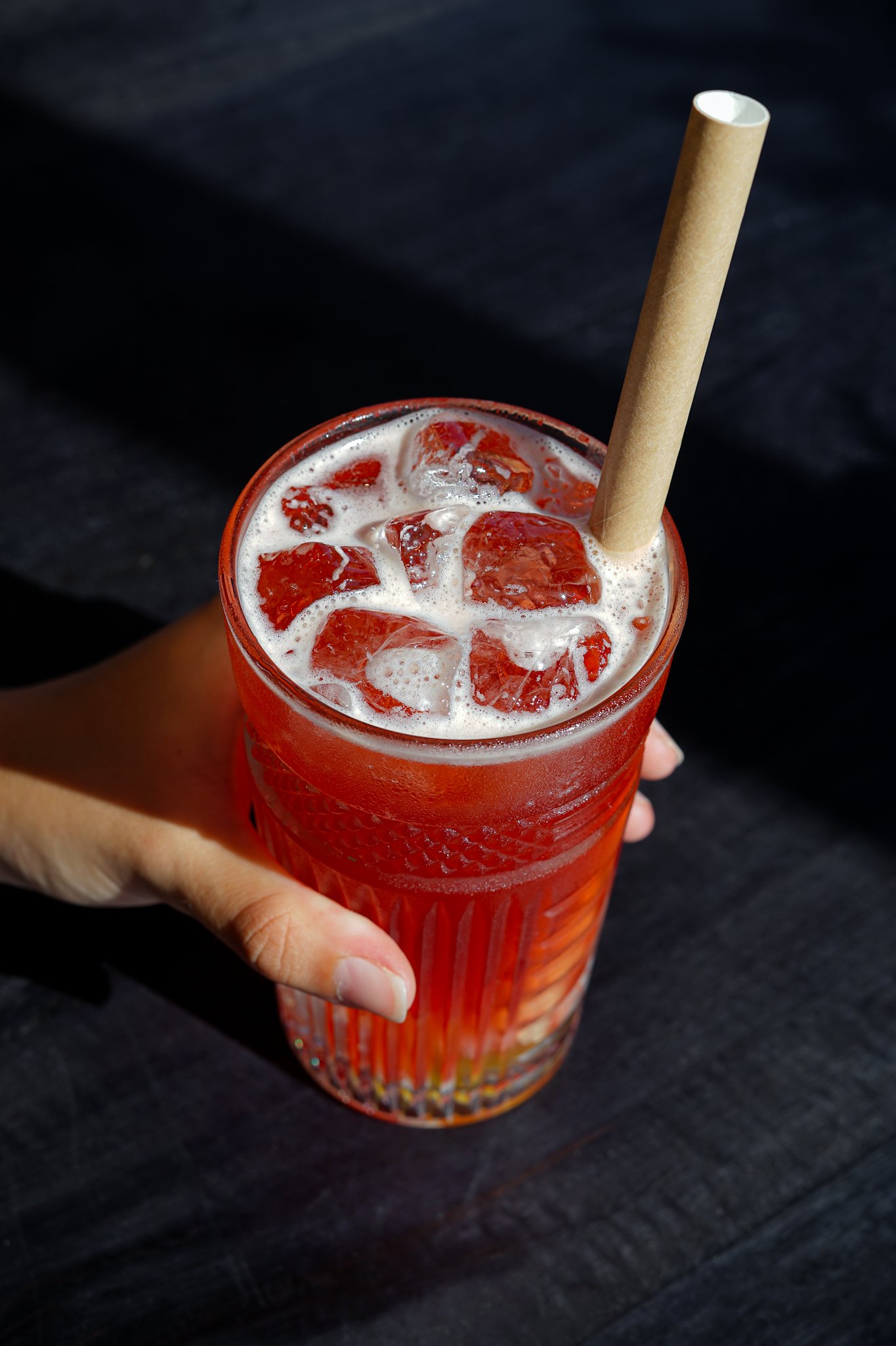 Verre rouge avec des glaçons et une boisson rouge, un gros tube en papier enrobe la boisson, tenu par une main sur fond noir.