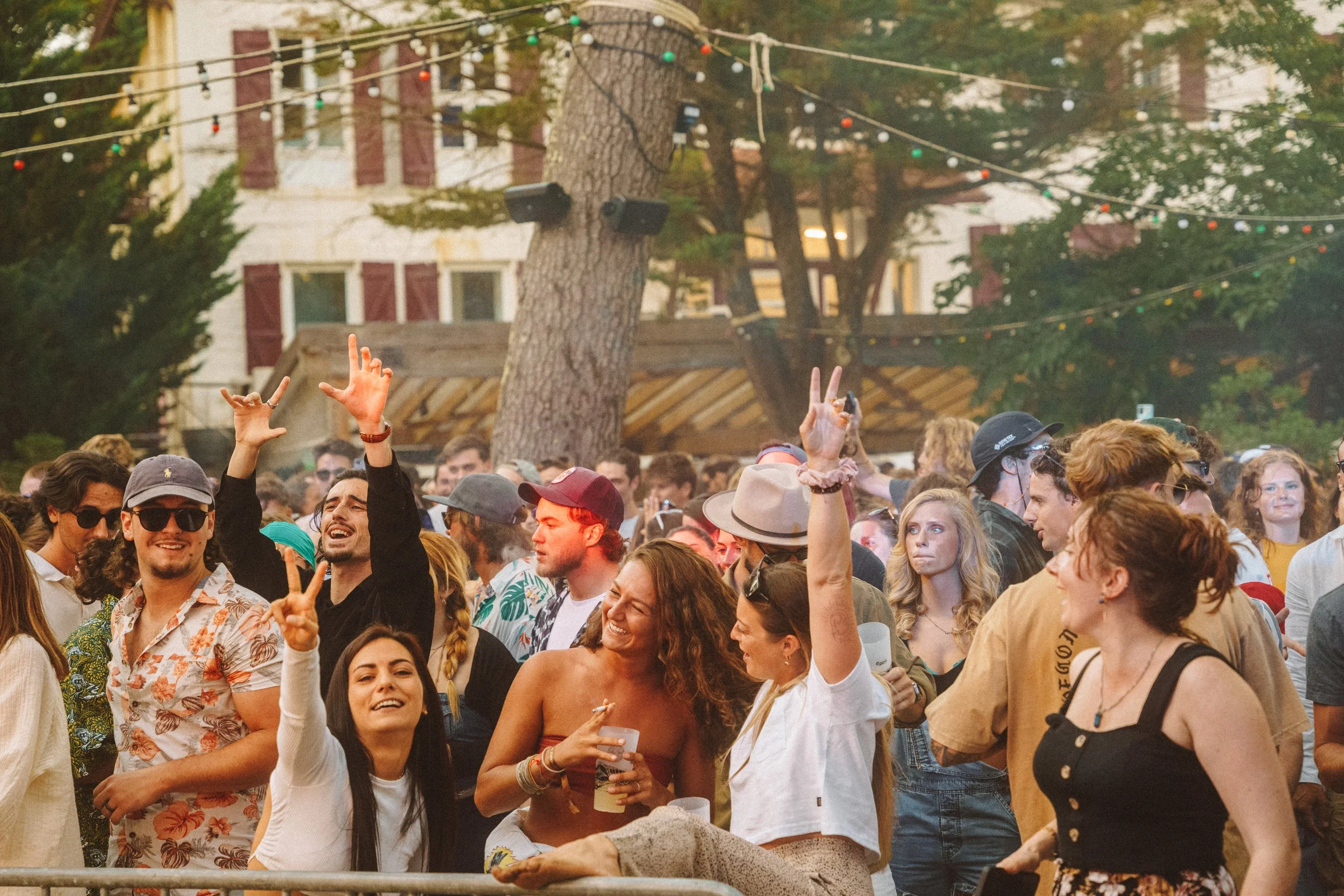 Un groupe de personnes souriantes et parlant lors d'un événement extérieur, les mains levées ou en mouvement, avec des arbres, des bâtiments et des guirlandes lumineuses en arrière-plan.