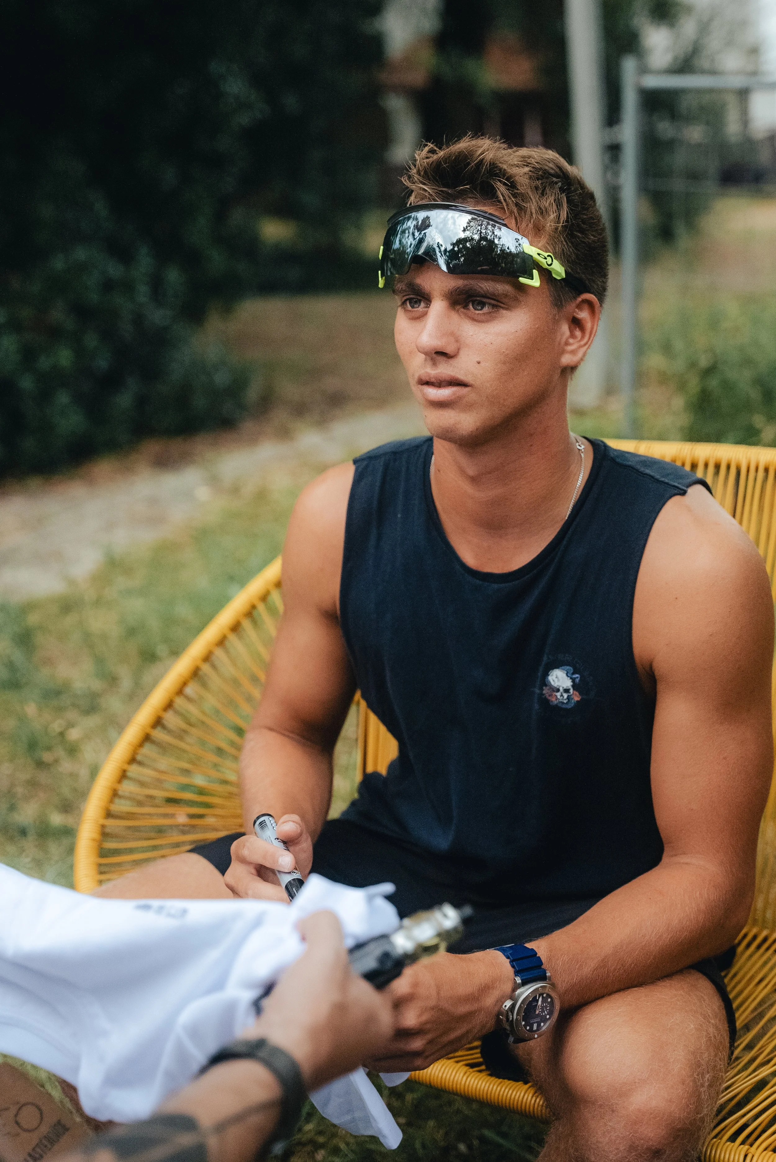 Un jeune homme avec des lunettes de soleil fumées, portant un t-shirt sans manches, est assis dans une chaise en rotin jaune à l'extérieur, en train de signer un autographe.