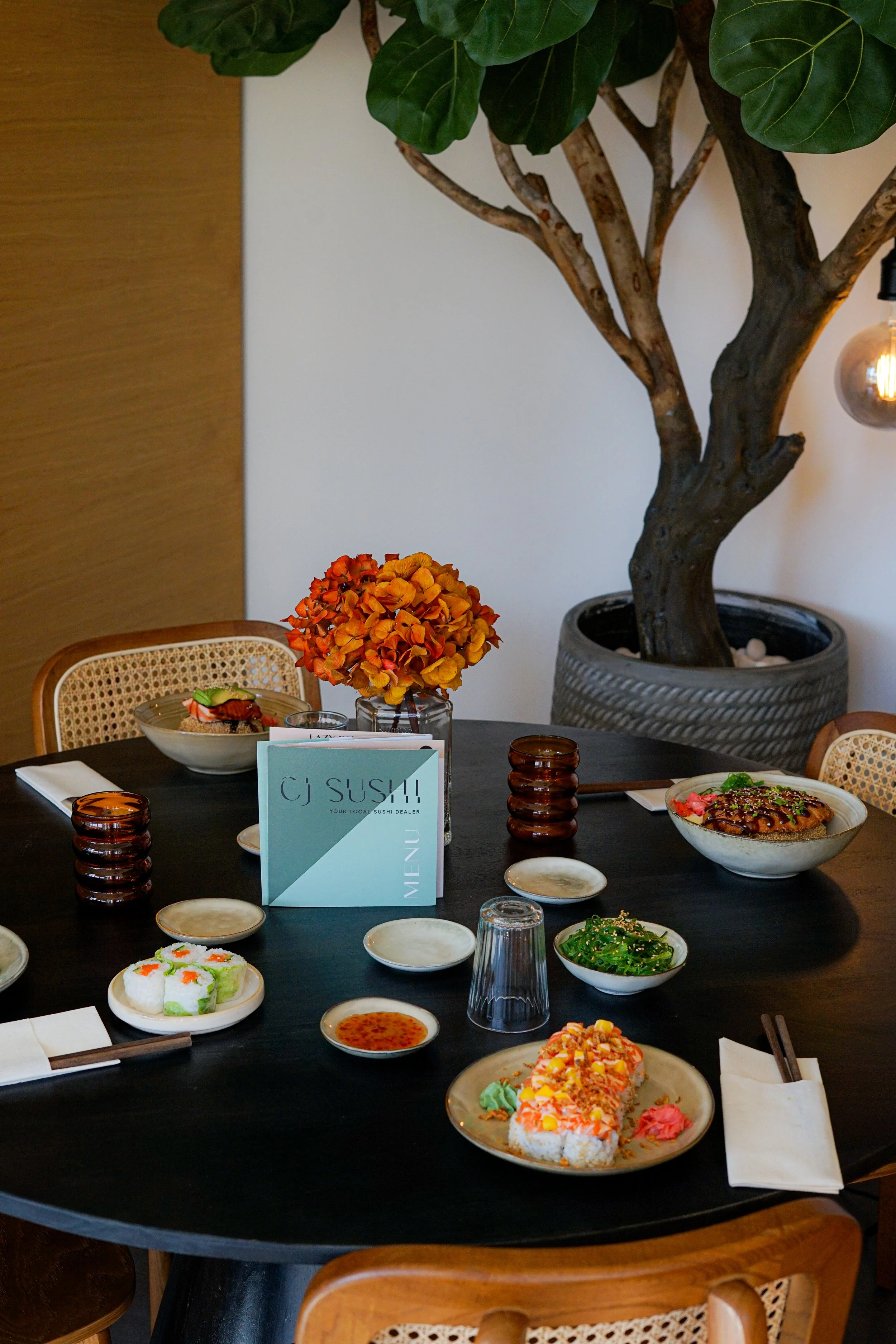 Une table dressée avec divers plats japonais, un menu de sushi, des assiettes de sushi, de la sauce, des feuilles de salade, des chopsticks, et des verres. En arrière-plan, un grand arbre d'intérieur et une décoration moderne avec une lumière suspend