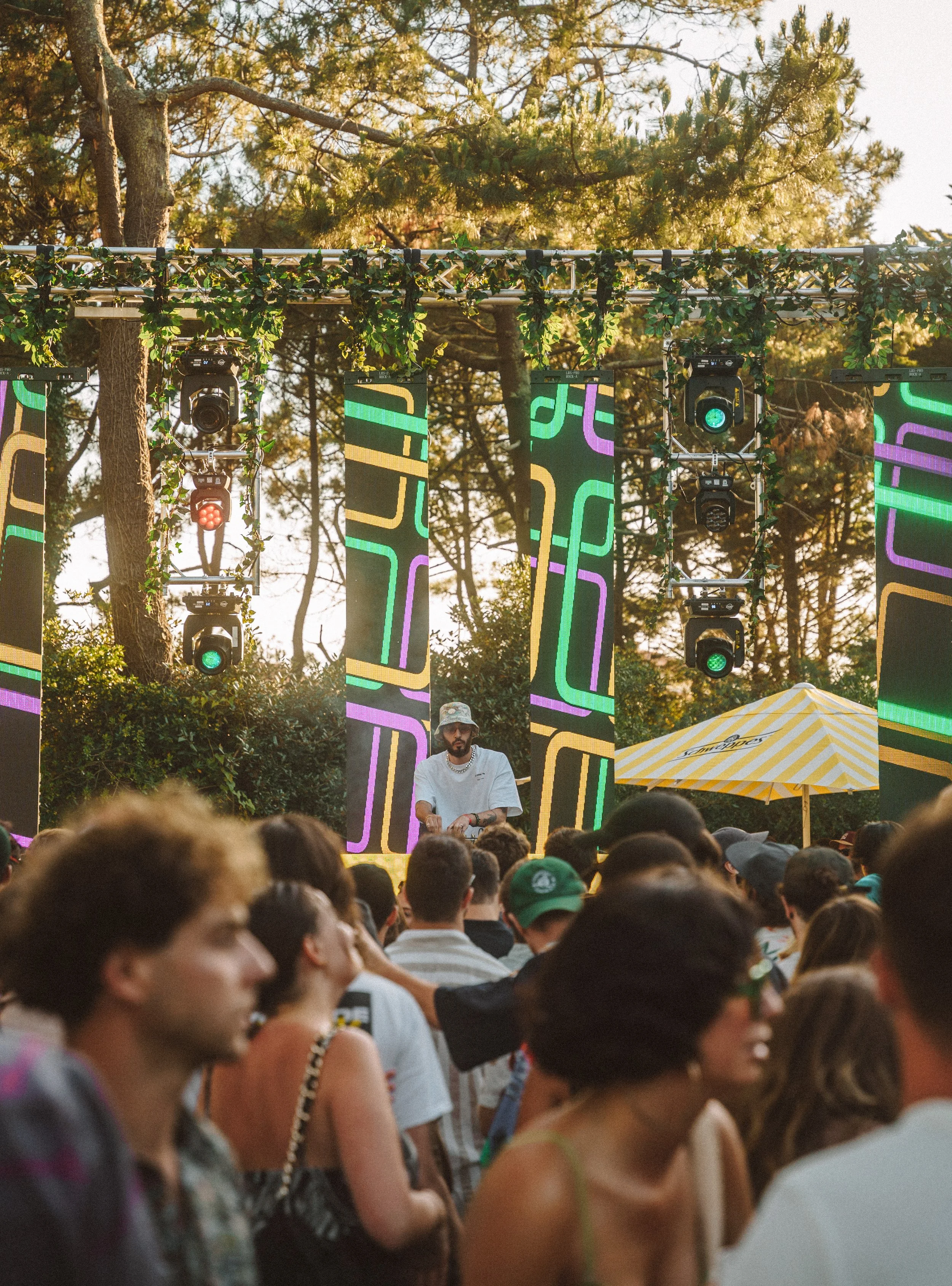 Un DJ joue de la musique lors d'un événement en plein air, avec un public nombreux, sous un ciel en soirée, entouré d'arbres, avec un système d'éclairage et des écrans LED colorés.