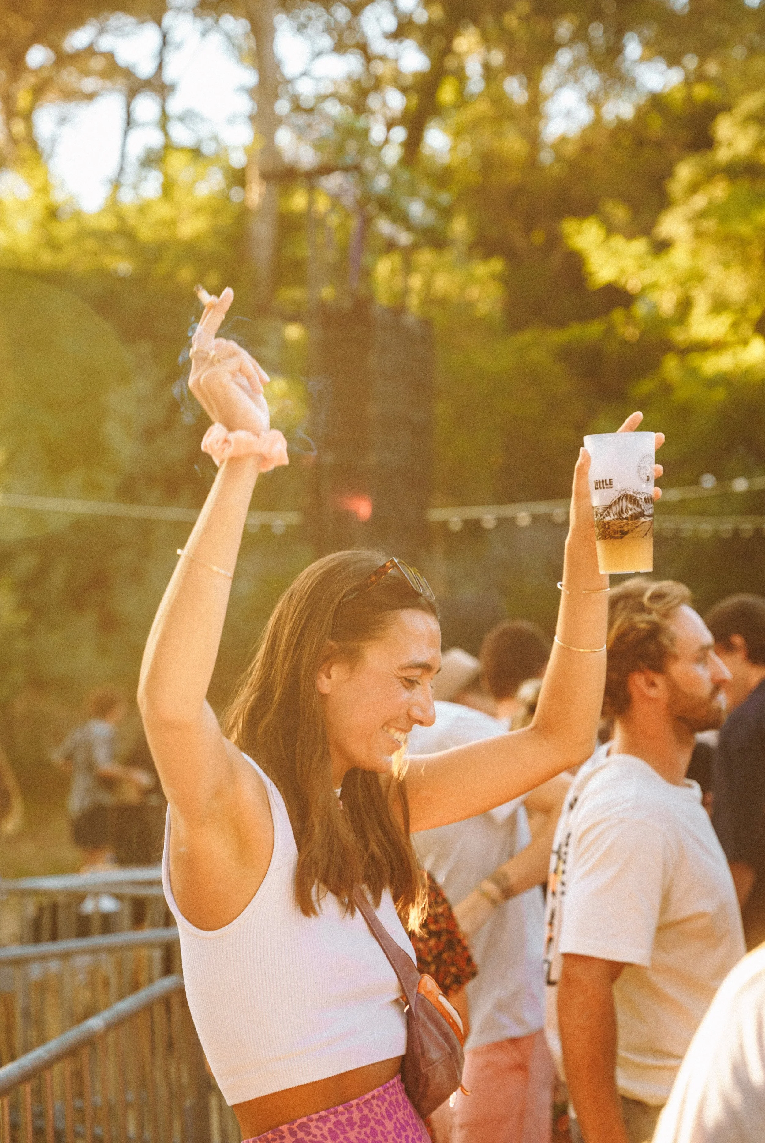 Une femme souriante lors d'un événement en plein air, levant un verre d'une main, avec d'autres personnes en arrière-plan, sous une lumière chaude de fin d'après-midi, entourée de nature.