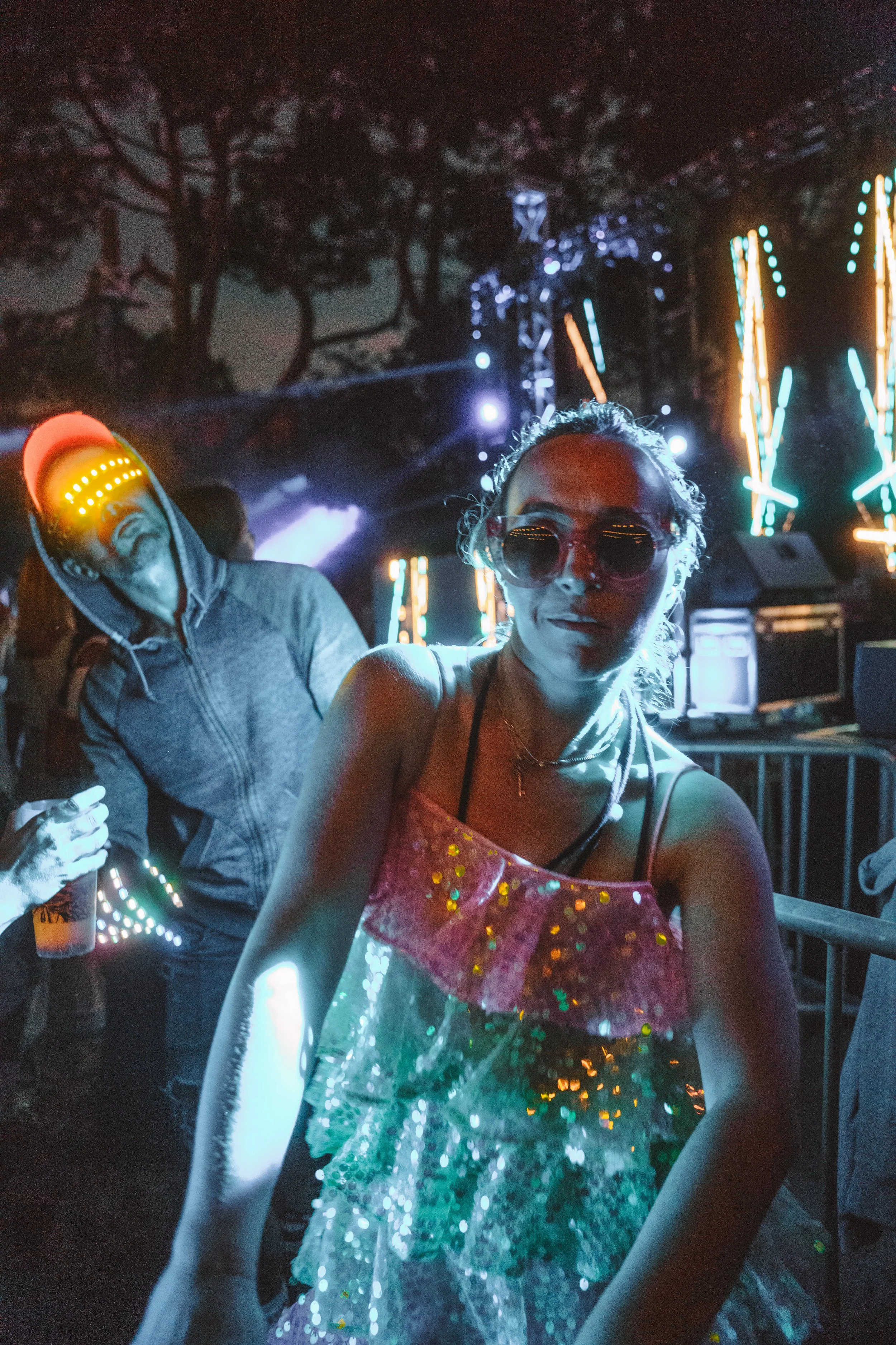 Une femme portant une robe à paillettes colorée danse dans une ambiance de fête avec des lumières vives et un décor nocturne, accompagnée d'un homme en sweat à capuche dans un lieu animé de nuit.