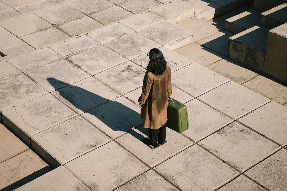 Une femme avec un manteau marron et des lunettes, tenant une valise verte, se trouve sur un pavement en béton, avec son ombre projetée sur le sol.