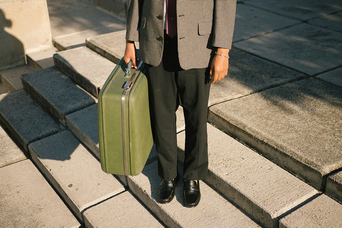 Person habillée en costume avec un valise verte à la main, marchant sur des marches en pierre, en plein jour.