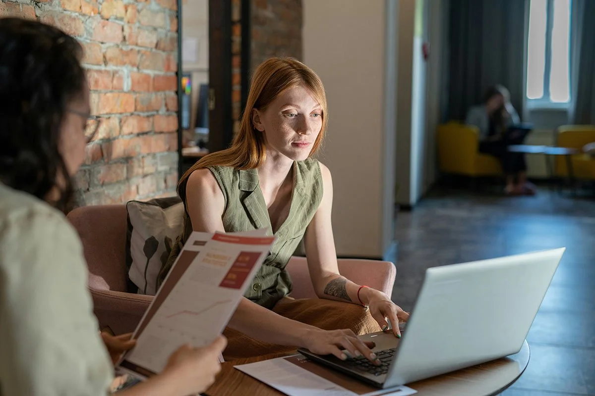 Deux femmes discutent dans un espace de travail, une à un ordinateur portable et l'autre avec des papiers.