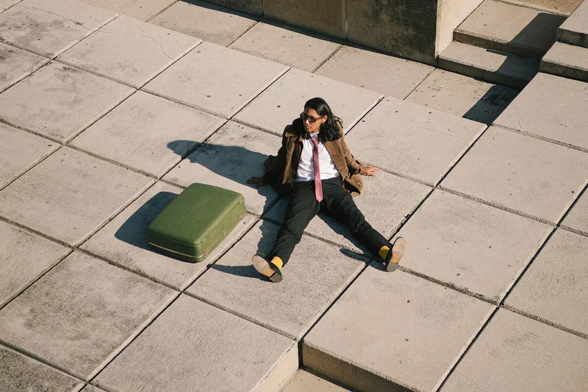 Une femme assise par terre sur un trottoir en pierre, portant des lunettes de soleil, une chemise blanche, une cravate rose, une veste marron et un pantalon noir, avec une valise verte à côté d'elle.