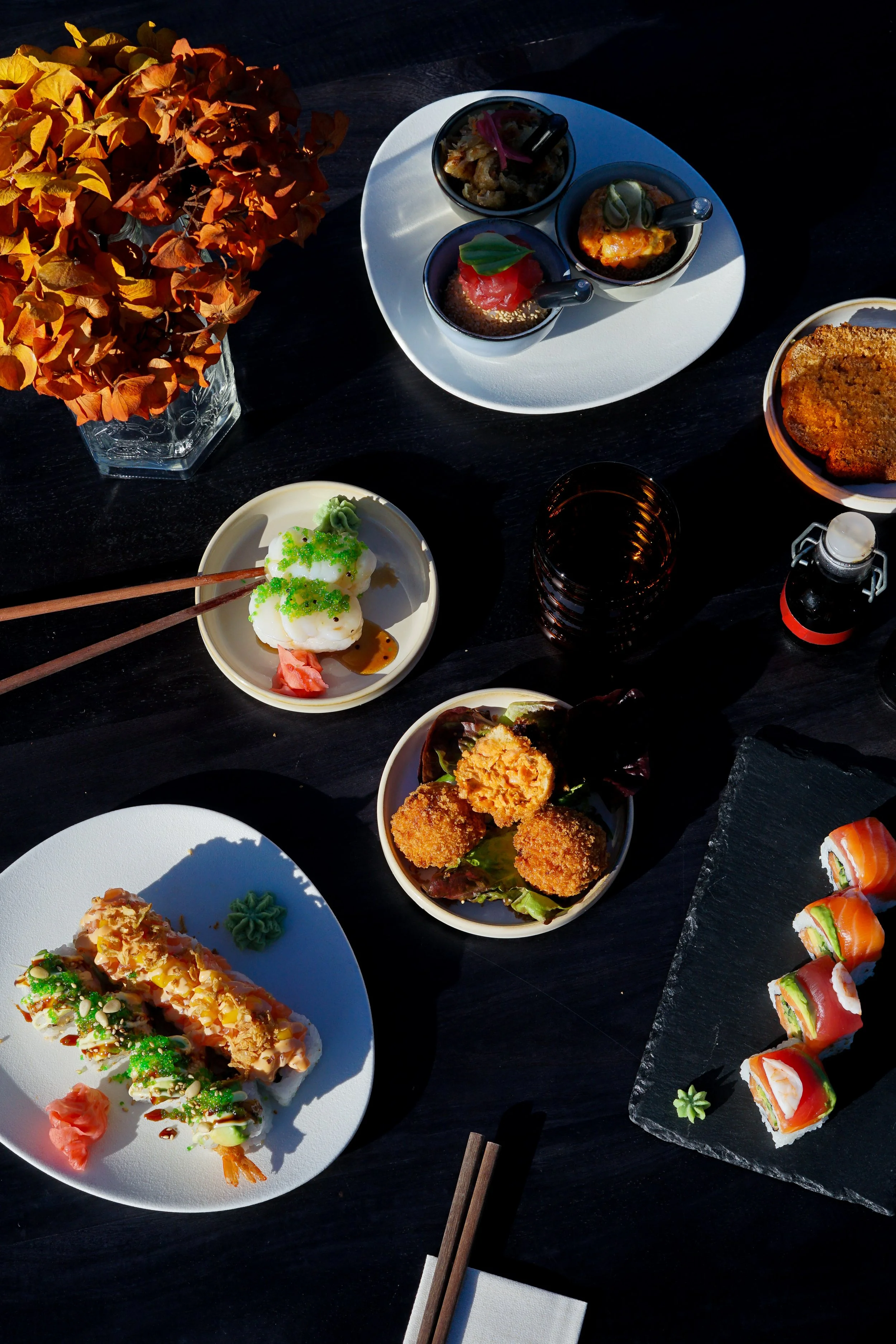 Assortiment de sushis, sashimis et autres plats japonais sur une table en bois noire, accompagnés d'une déco florale orange.