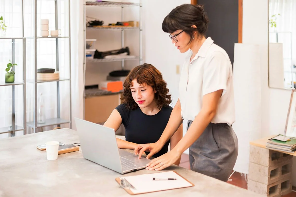 Une femme montrant quelque chose à un ordinateur portable à une autre femme dans un bureau lumineux.