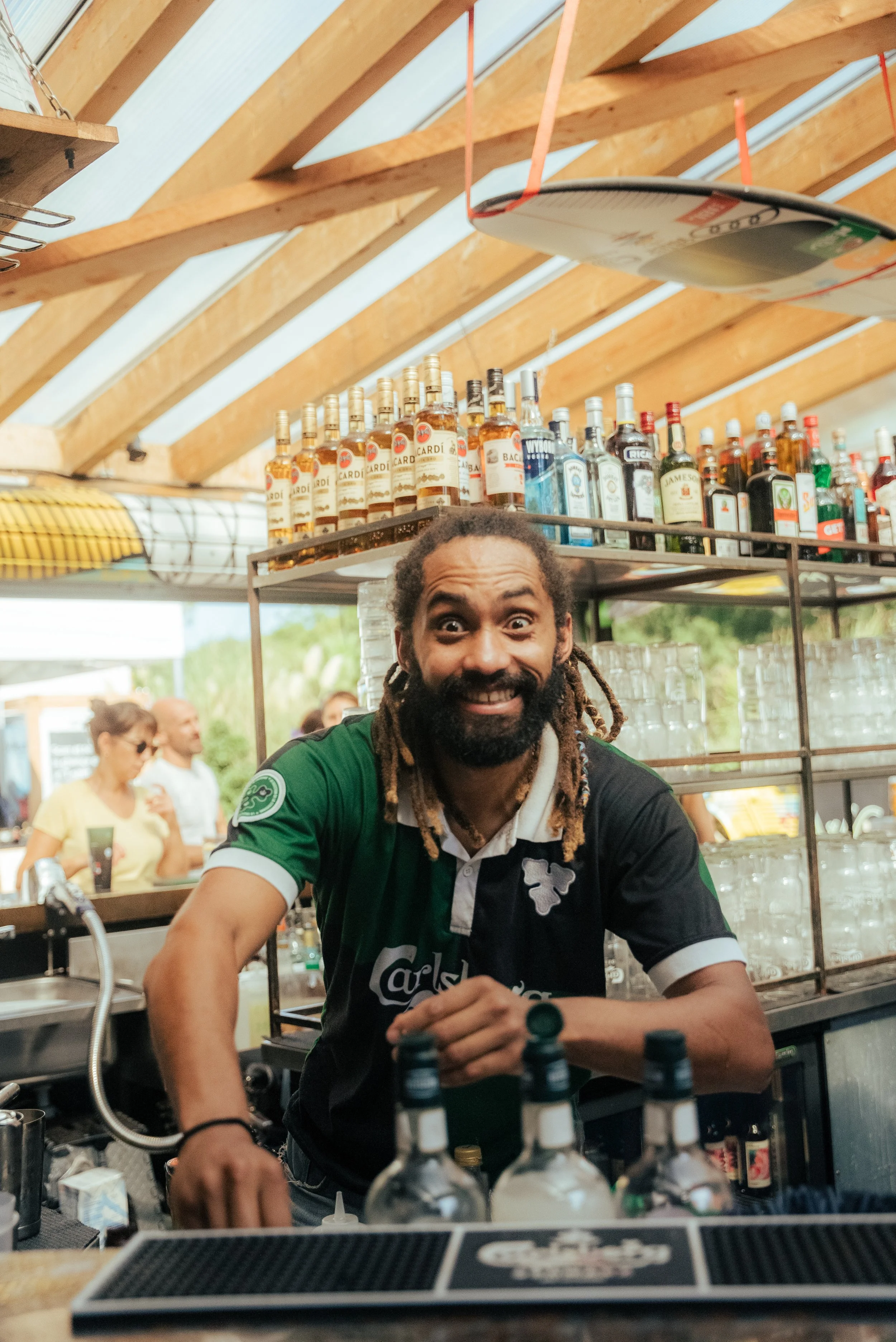 Homme souriant derrière un bar avec des bouteilles d'alcool, portant un maillot de football noir et vert Carlsberg, dans un endroit animé, probablement un bar ou un restaurant.