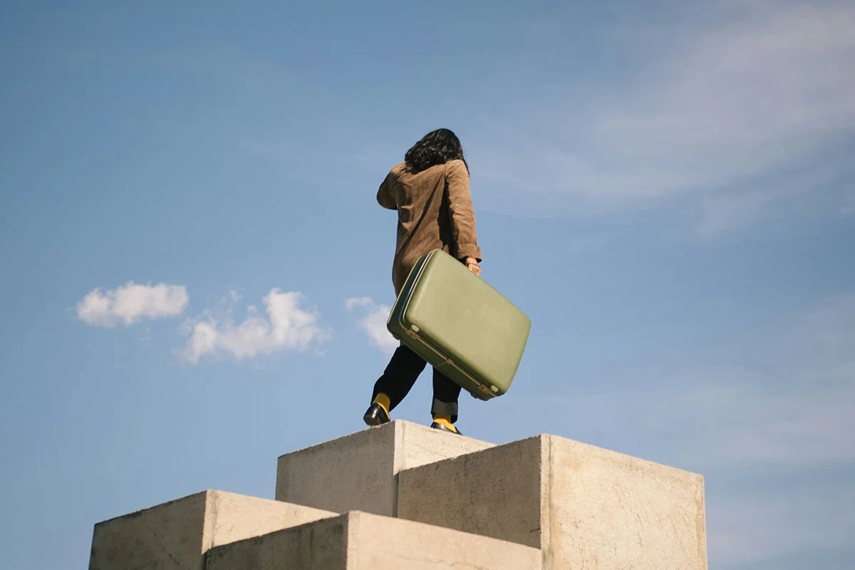 Une femme vue de dos qui se tient sur un cube en béton, tenant une valise verte, sous un ciel bleu avec quelques nuages.