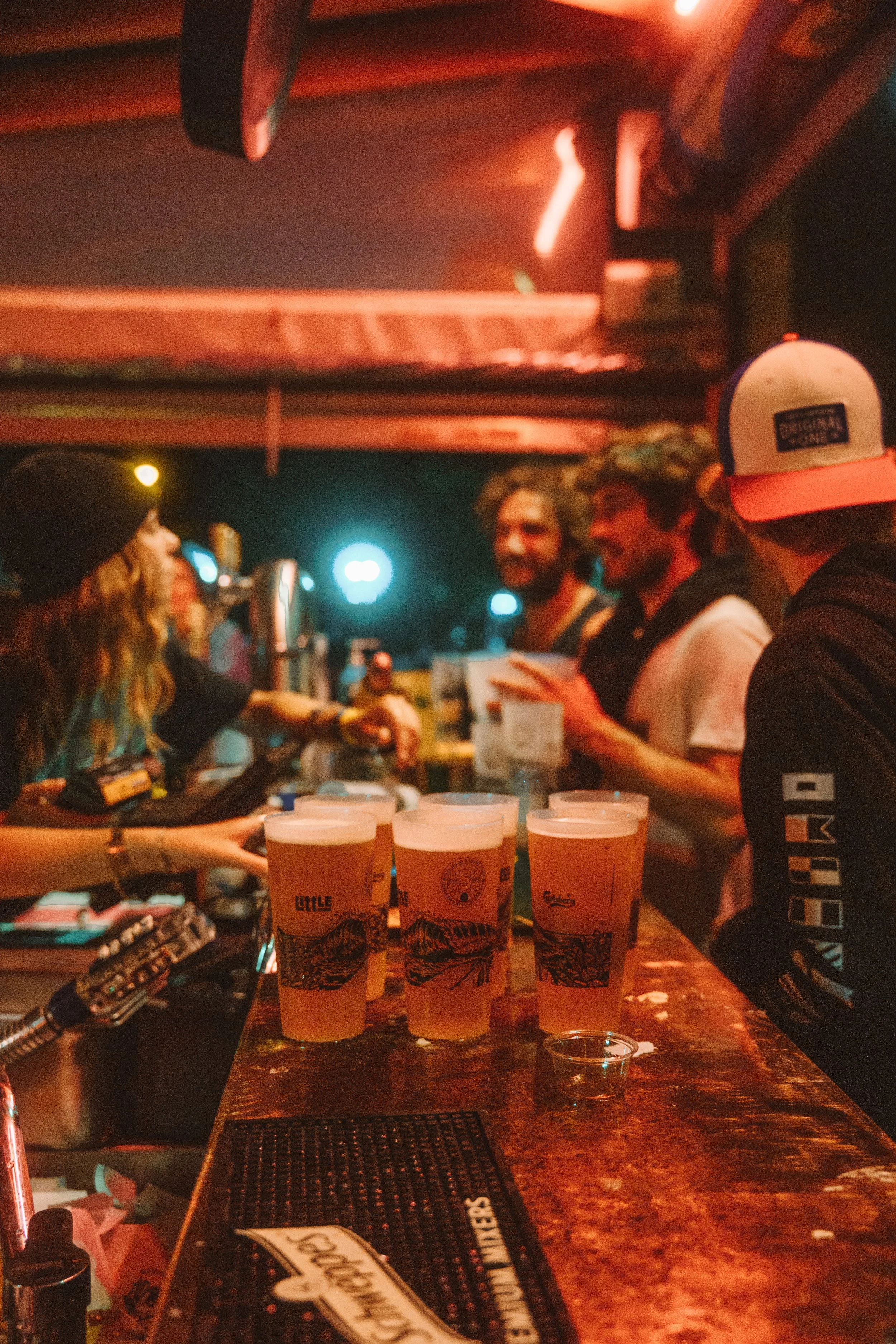 Verre de bière blonde sur un comptoir en bois dans un bar avec plusieurs personnes discutant en arrière-plan, ambiance chaleureuse et intimiste.