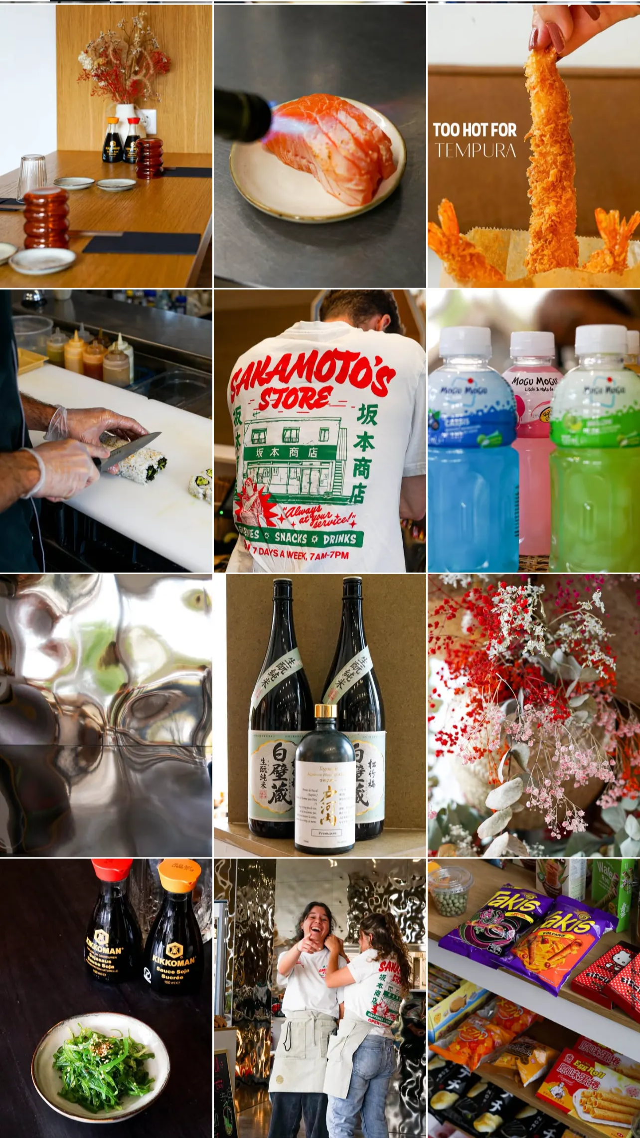 Une table de restaurant avec des assiettes, des condiments et des fleurs, une assiette de sashimi, une friture de tempura, un robot manuel en train de faire des sushis, une affiche de Sakura's Store, des bouteilles de sauces soja, des bouteilles d'alcool japonais, un bouquet de fleurs, des bouteilles de sauce soja, deux jeunes femmes riant en uniforme de restaurant, des étagères avec des snacks japonais.