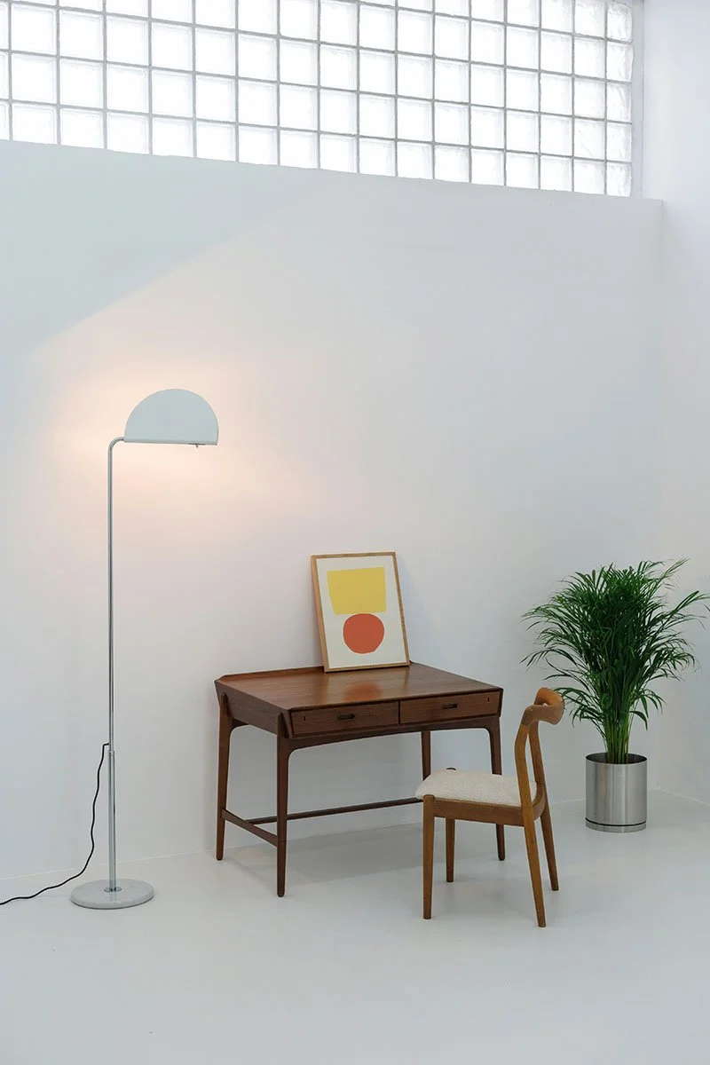 Un intérieur minimaliste avec un mur blanc, une lampe sur pied, une table en bois avec un tableau abstrait dessus, une chaise en bois avec un coussin et un grand pot avec une plante verte.