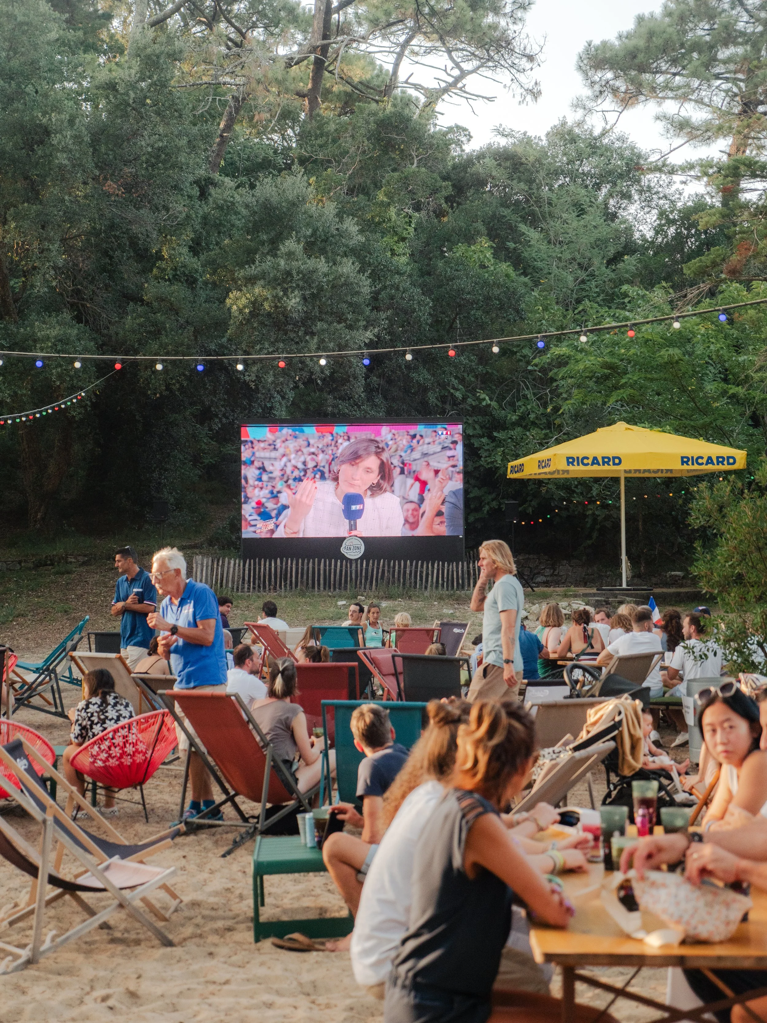 Une fête en plein air avec des personnes assises sur des chaises et des tables, un grand écran diffusant une interview, une grande parasol jaune avec la marque Ricard, des guirlandes lumineuses suspendues entre les arbres, et une forêt dense en arriè