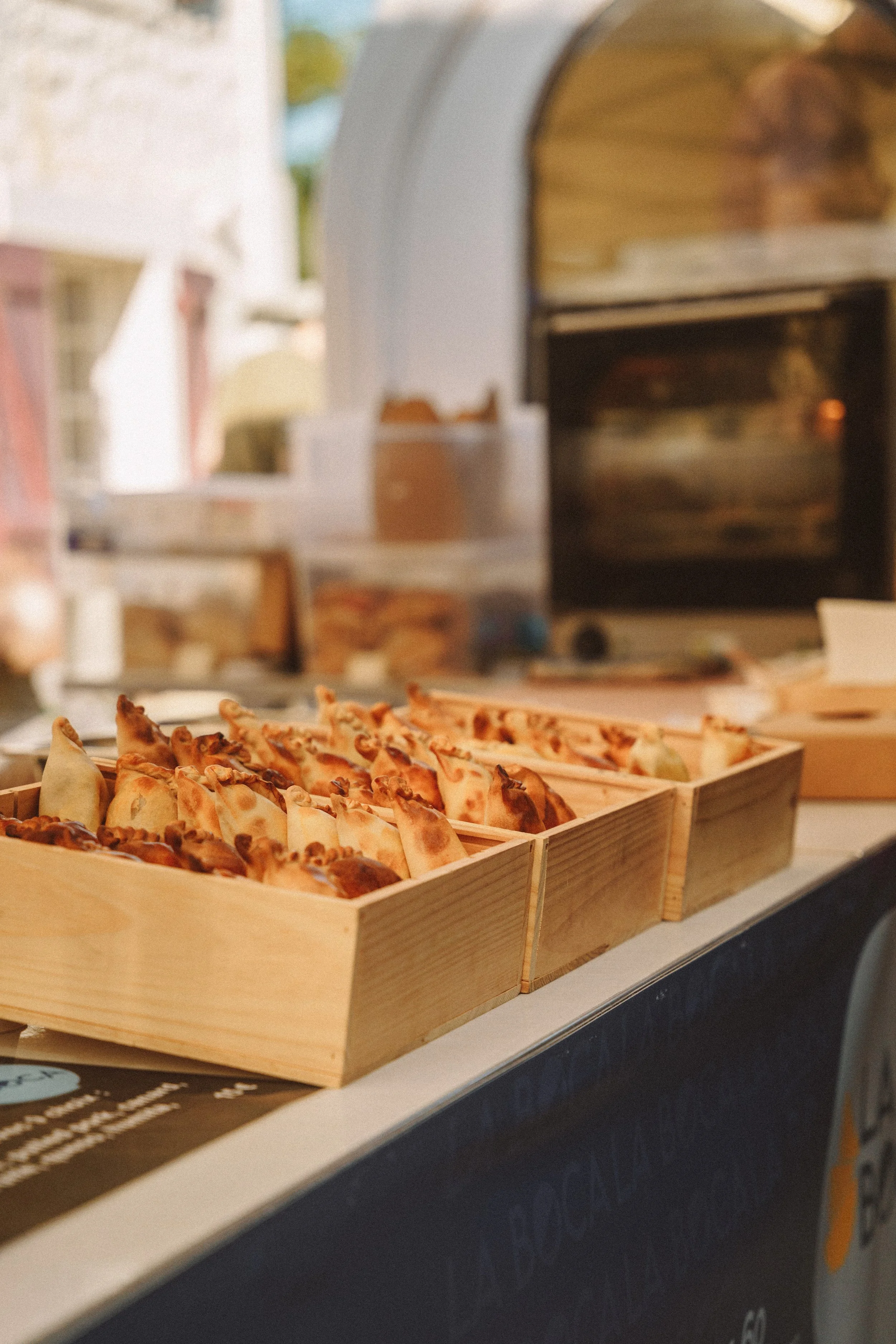 Plateau de petites pizzas ou quiches dans une boîte en bois, avec un four à pizza ou un four à bois en arrière-plan.