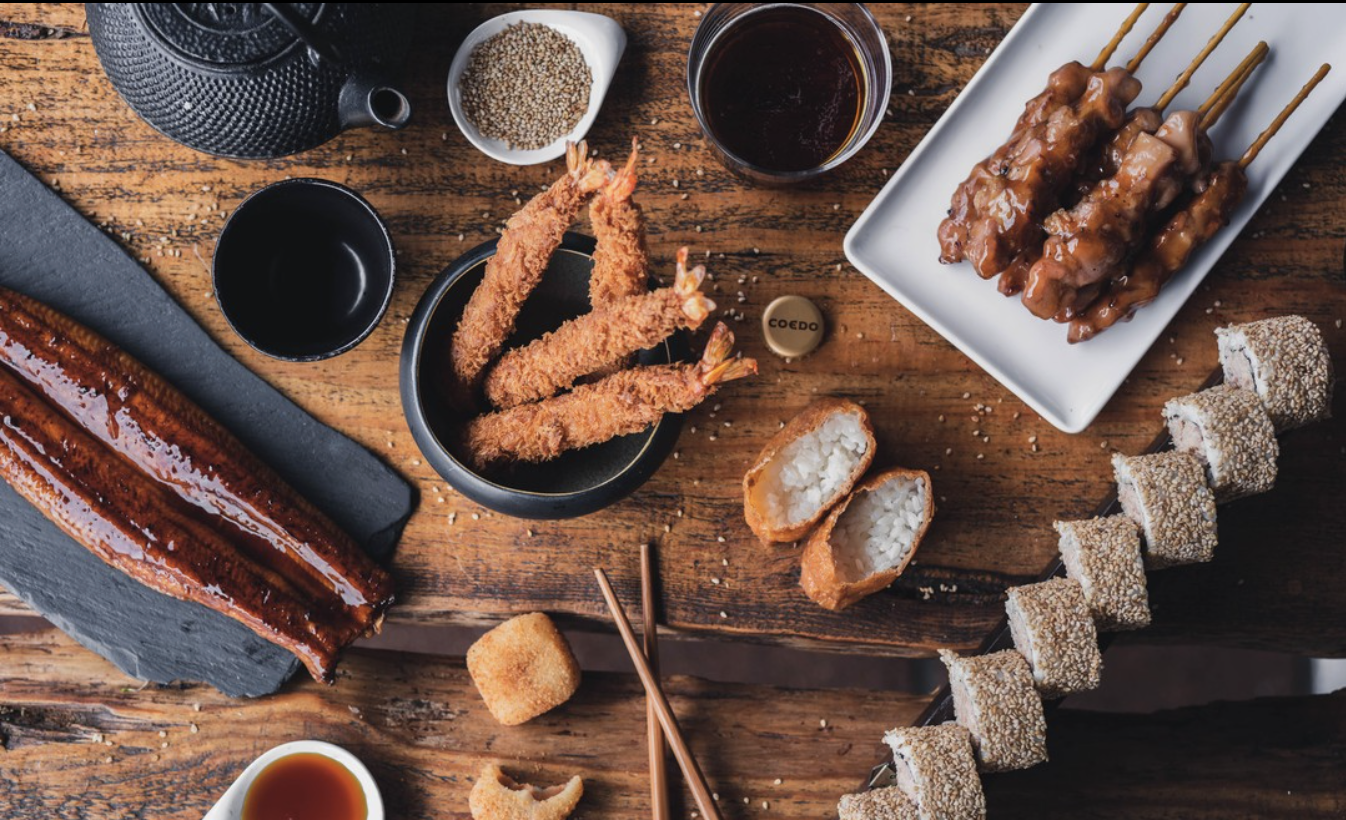 Variété de plats asiatiques, notamment des crevettes frites, des brochettes de viande, du sushi en rouleaux, un grand morceau de poisson grillé, ainsi que des sauces et condiments sur une table en bois.