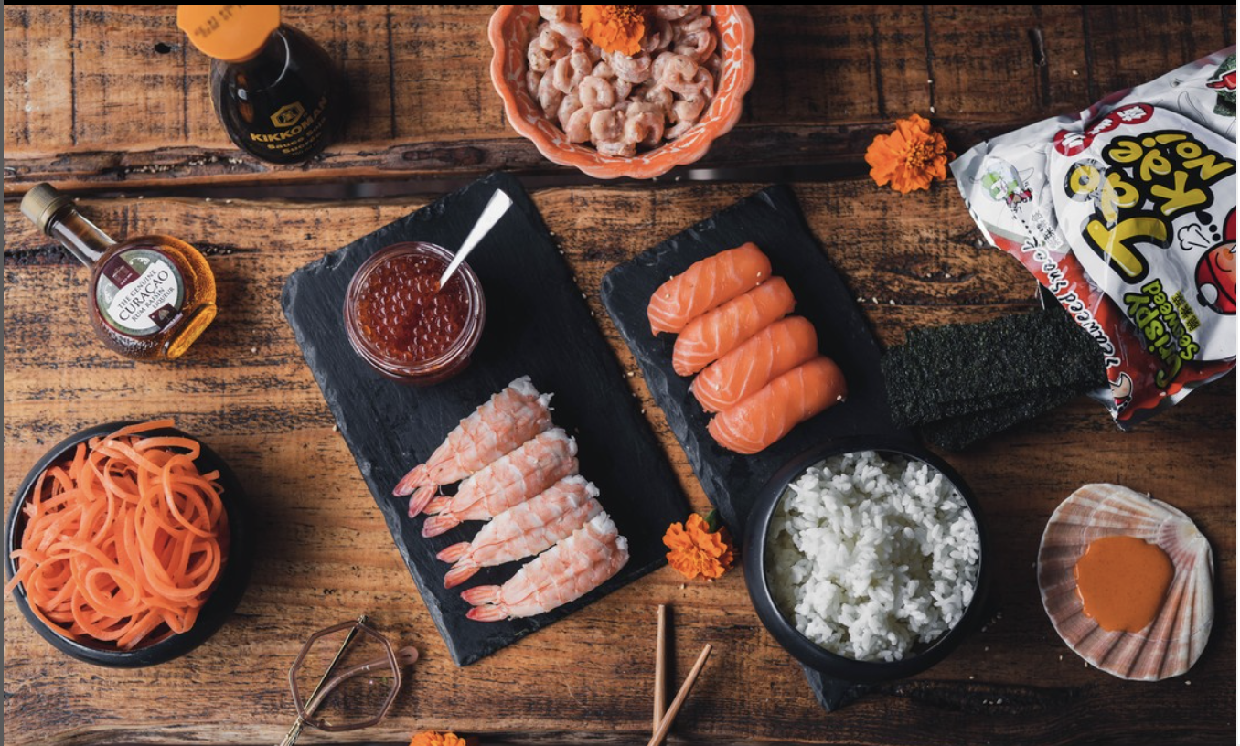 Ingrédients pour sushi sur une table en bois, incluant du riz, saumon, crevettes, algues nori, poisson en sauce, sauces, et snacks.