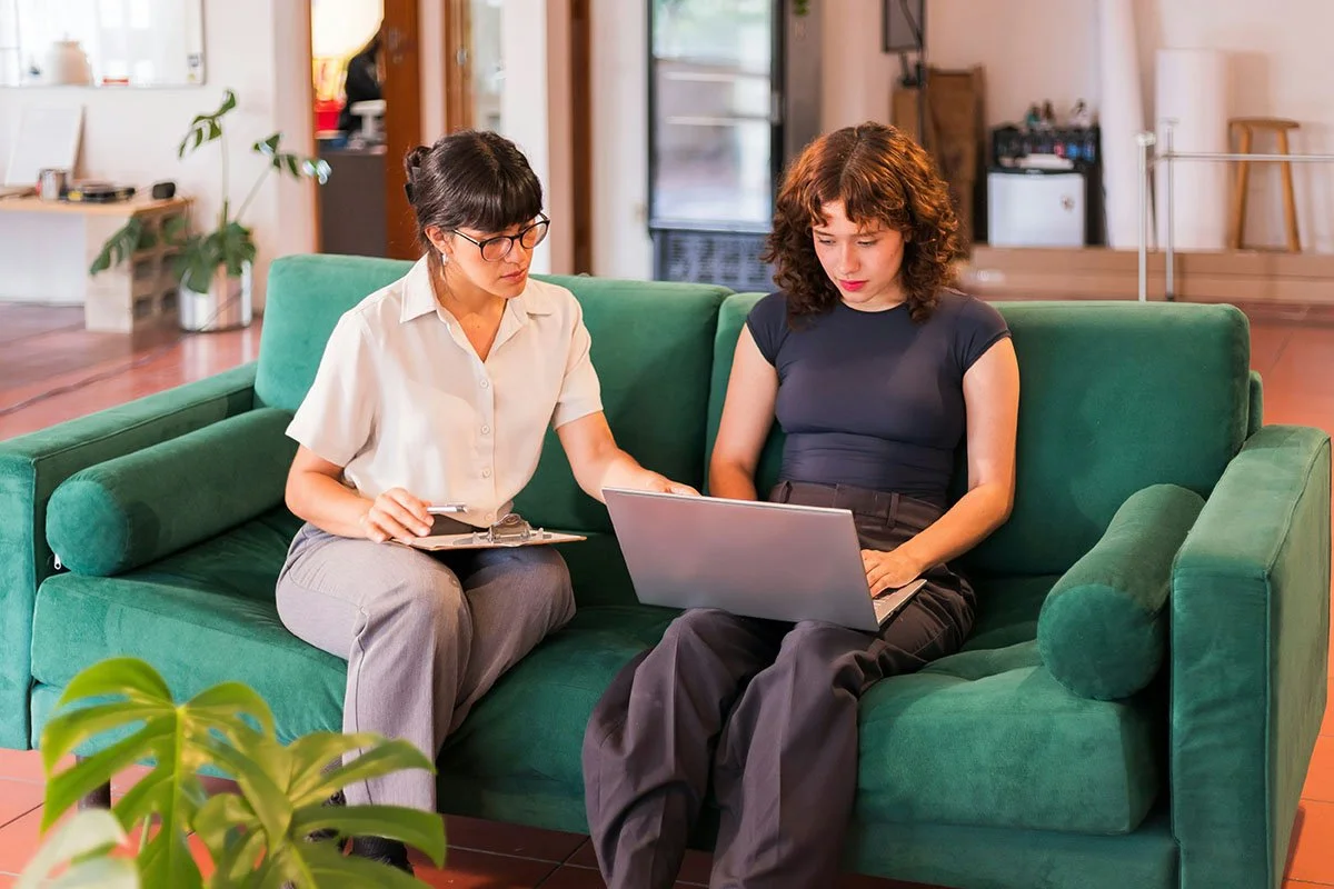 Deux femmes assises sur un canapé vert, discutant et regardant un ordinateur portable.