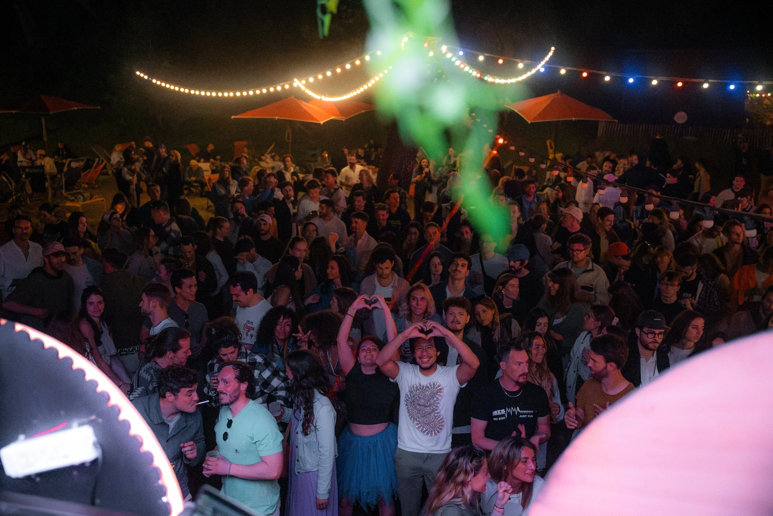 Une foule de personnes assises et debout à une fête nocturne avec des lumières colorées suspendues, des parasols orange et des décorations lumineuses.