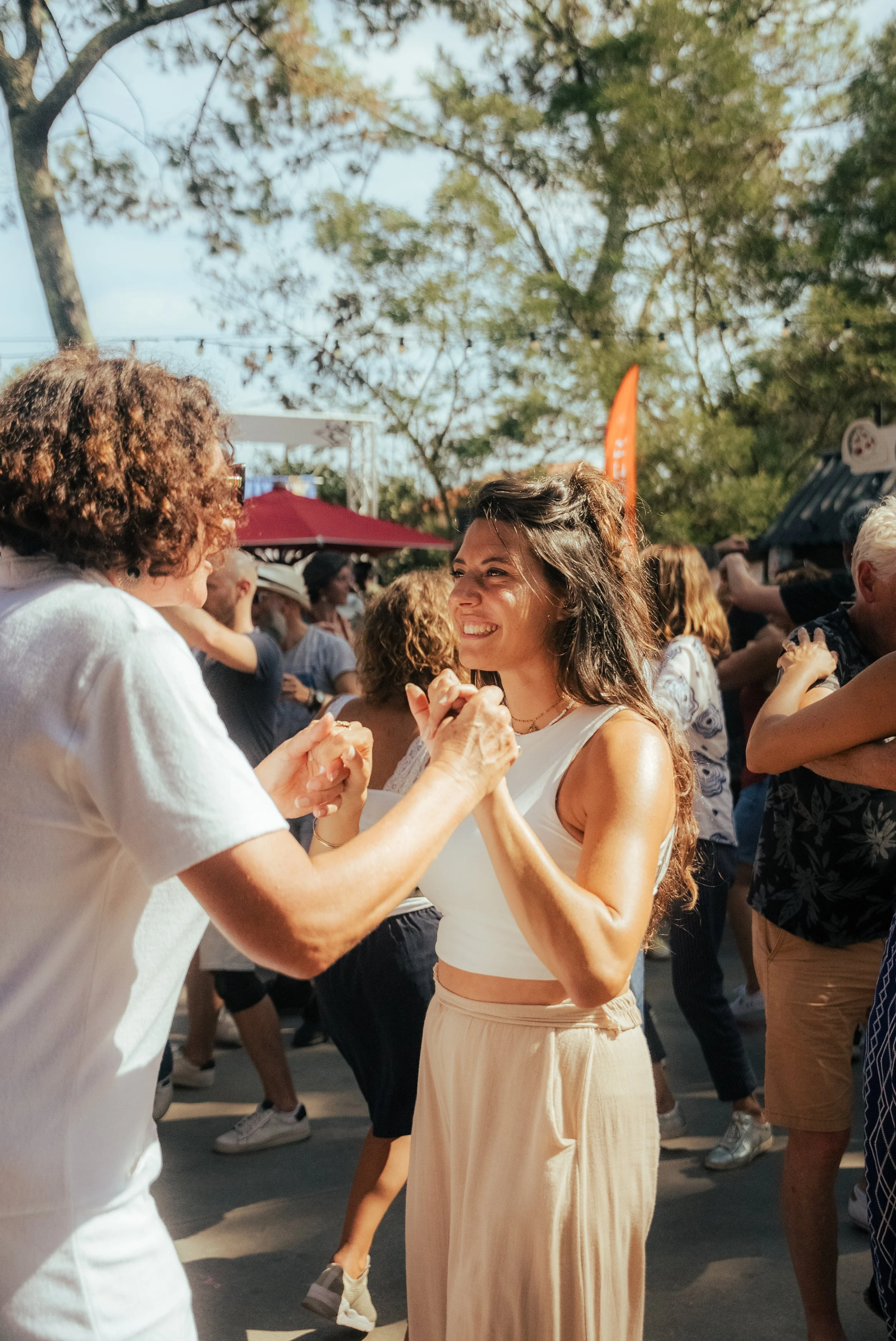 Deux femmes dansent ensemble lors d'un événement en plein air, entourées d'autres personnes, avec des arbres en arrière-plan.