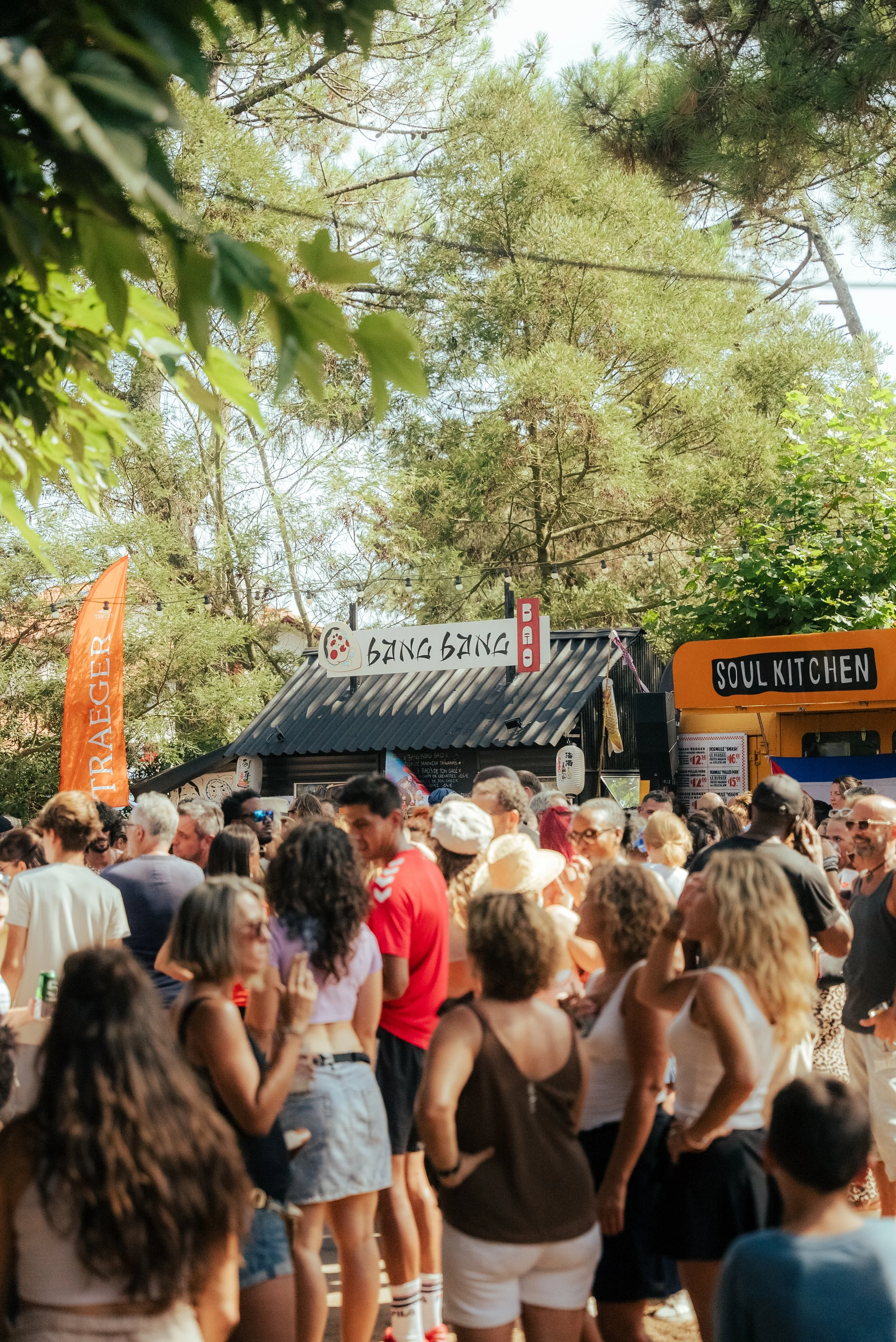 Une foule de personnes debout dans un festival en plein air sous un ciel ensoleillé, avec des arbres verts en arrière-plan. Il y a des stands de nourriture ou de boissons, dont un avec une enseigne 'Soul Kitchen' et un autre avec 'Bang Bang'.