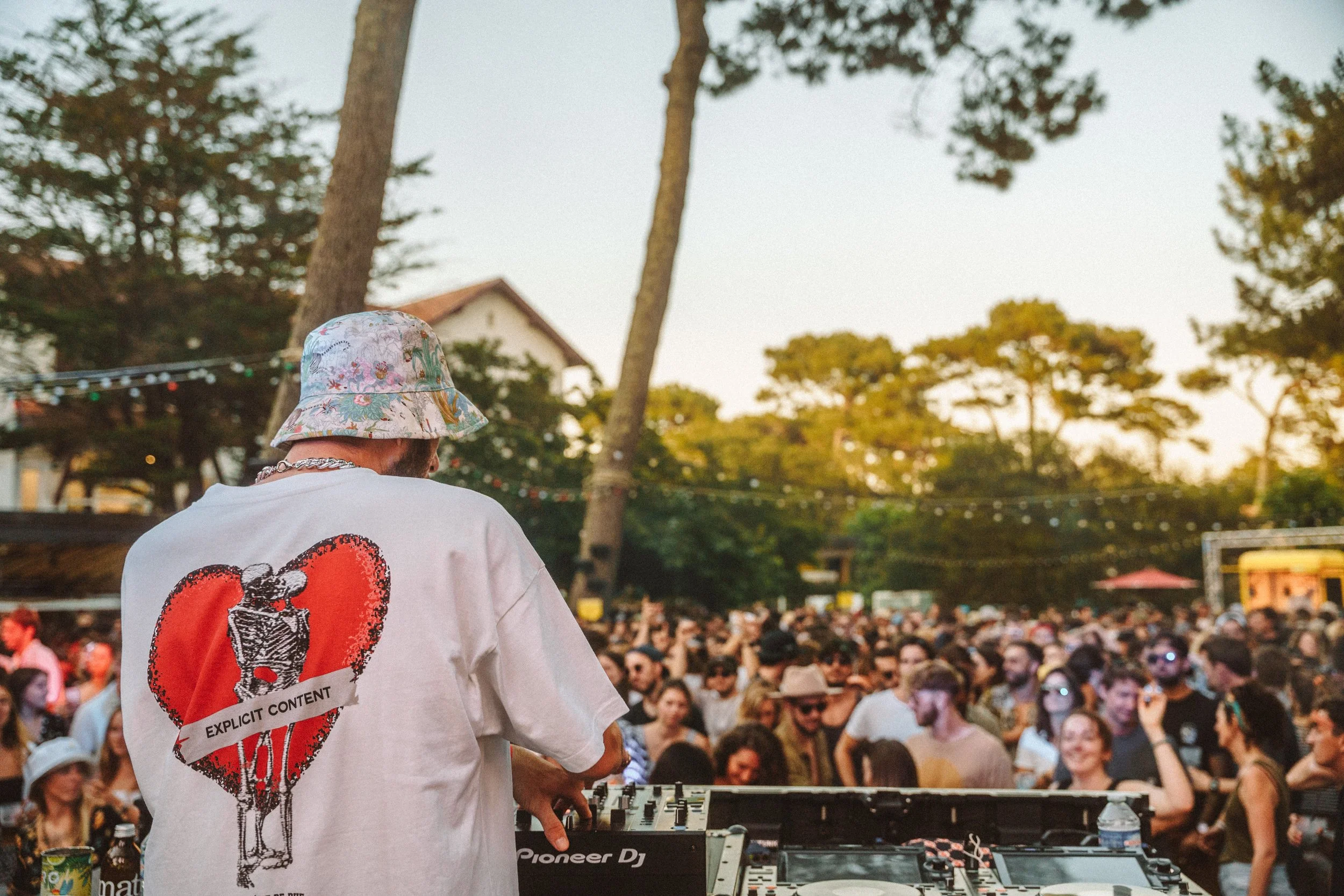 Une personne portant un chapeau à fleurs et un T-shirt avec un cœur rouge, un bretzel et la phrase 'Explicit Content', est DJ lors d'un grand rassemblement en plein air avec une foule et des arbres en arrière-plan.