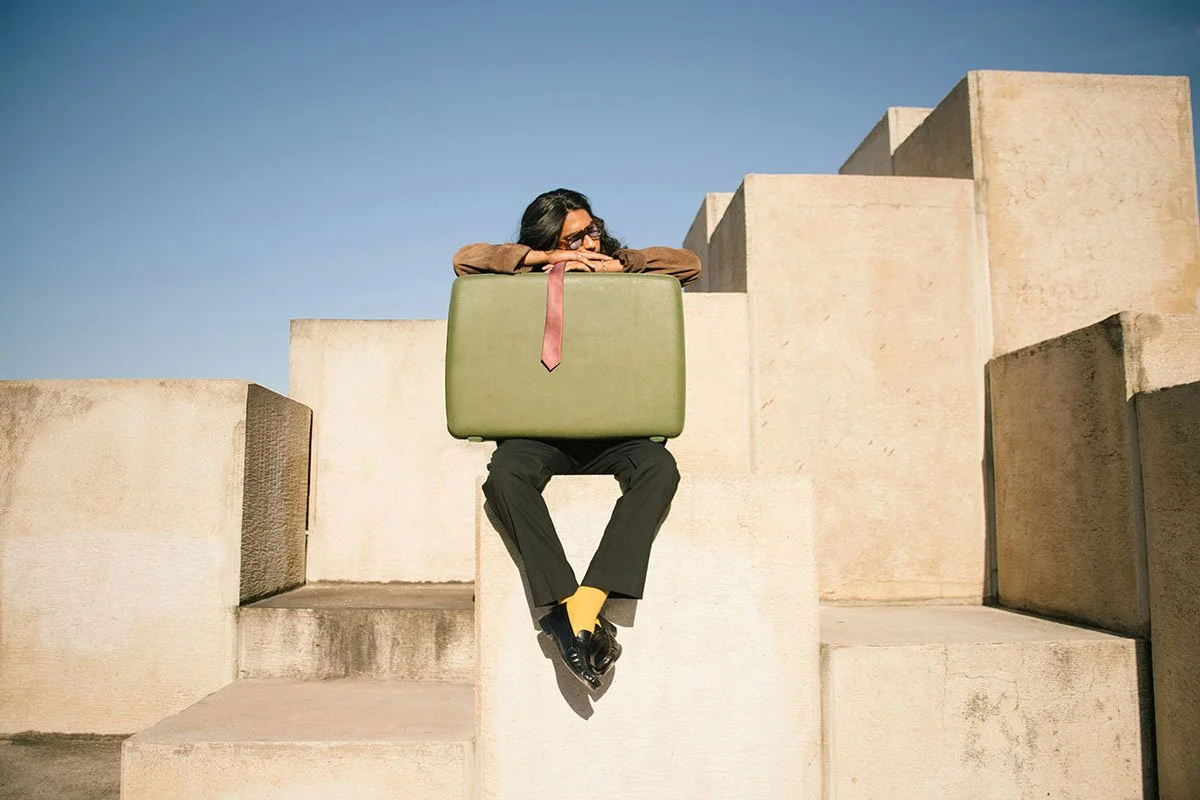 Une femme assise sur une structure en béton, portant une grande valise verte sur ses genoux, regardant pensivement.