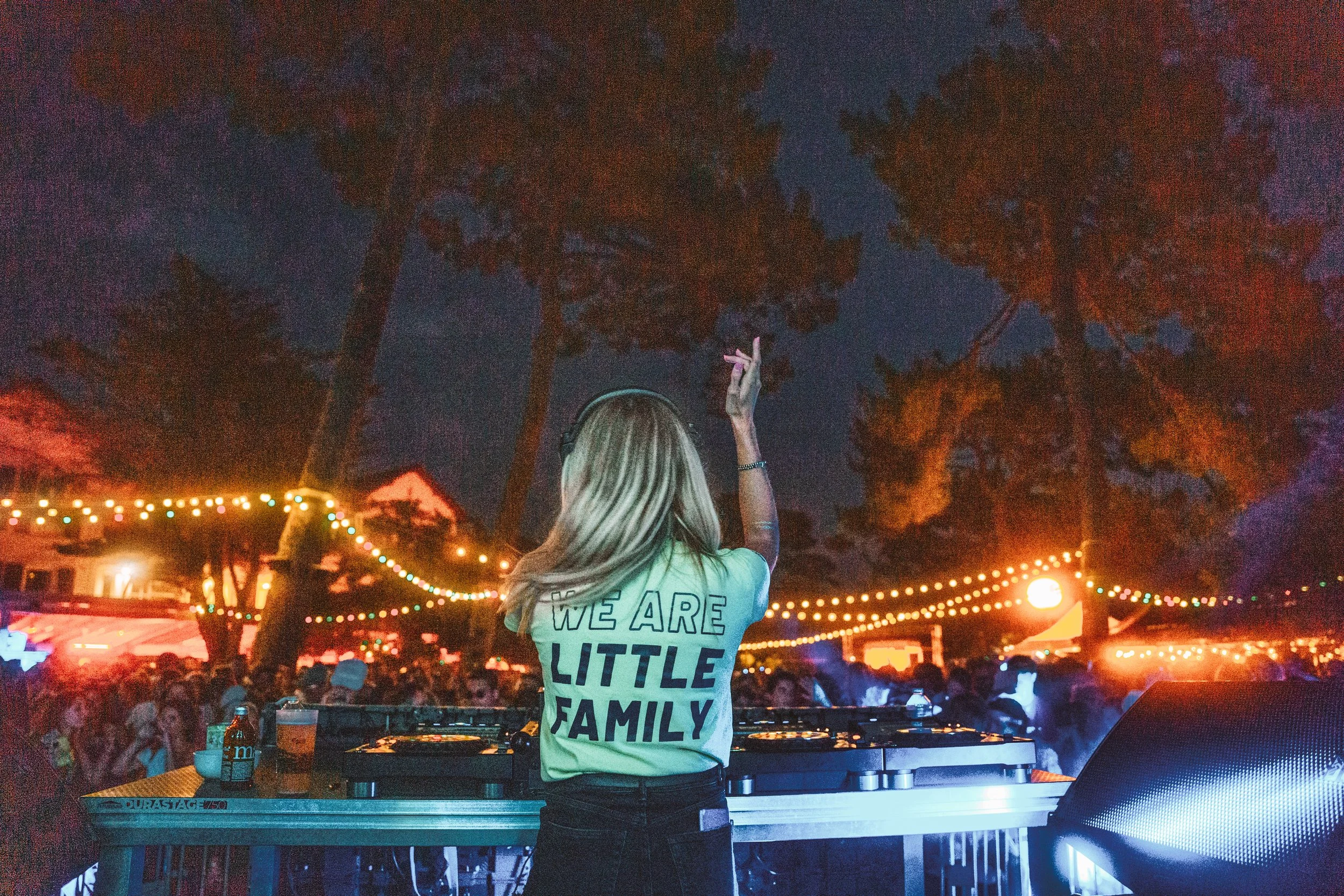 Une femme de dos portant un t-shirt avec l'inscription « we are a little family » danse ou agit devant un public lors d'un événement nocturne avec des lumières décoratives et des arbres en arrière-plan.