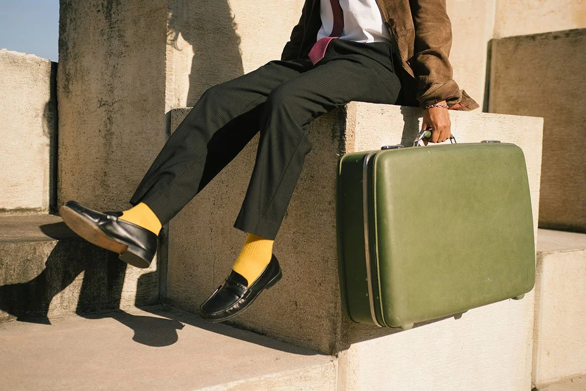 Une personne assise sur un bloc de pierre avec une valise verte à côté, portant des pantalons noirs, une chemise blanche, une veste marron, des chaussettes jaunes et des chaussures noires.