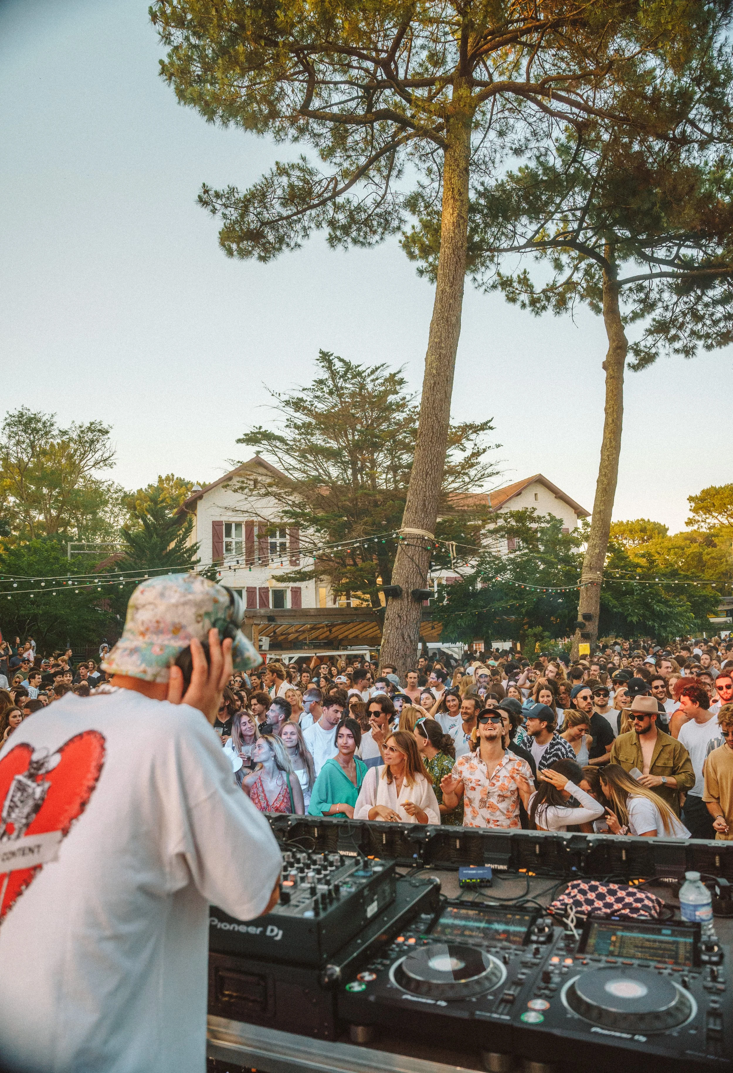 Un DJ joue de la musique devant une grande foule lors d'un festival en plein air par une fin d'après-midi ou début de soirée, avec des arbres et un bâtiment résidentiel en arrière-plan.