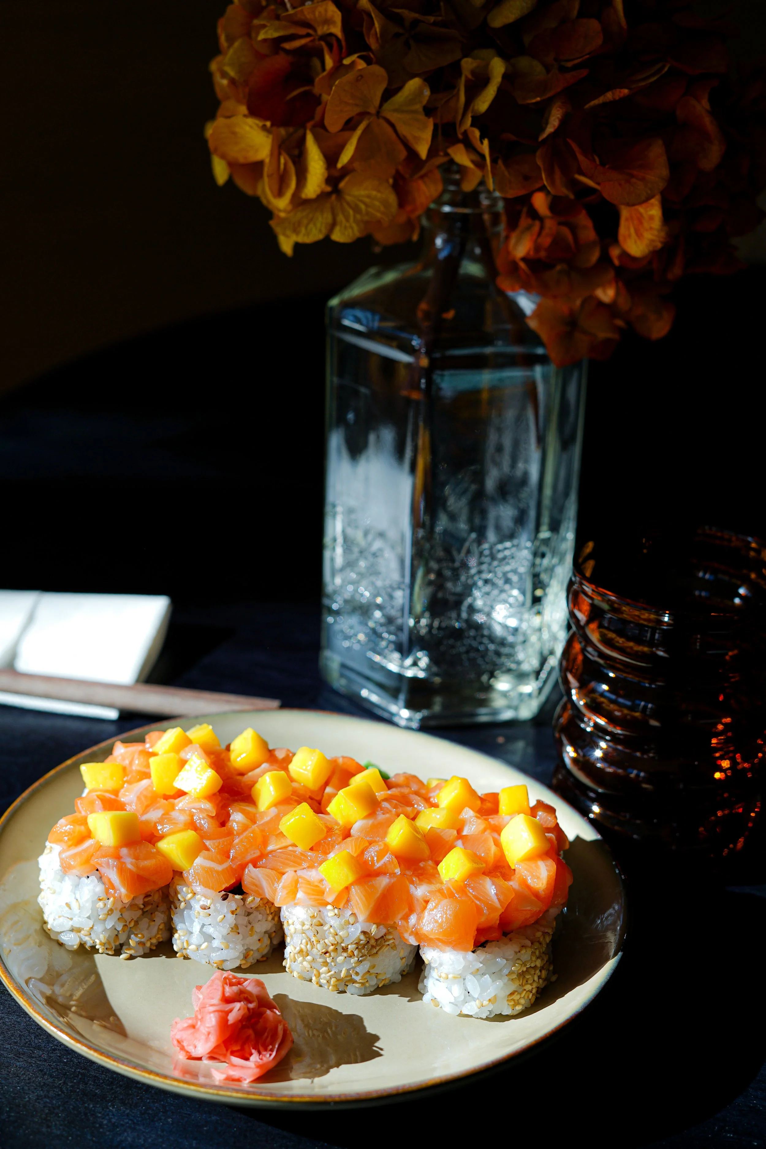 Un plat de sushi avec du saumon, de la mangue, et du riz, accompagné de wasabi, sur une assiette beige. En arrière-plan, une bouteille en verre avec des fleurs séchées, et un verre en verre foncé. La scène est illuminée par une lumière naturelle fort