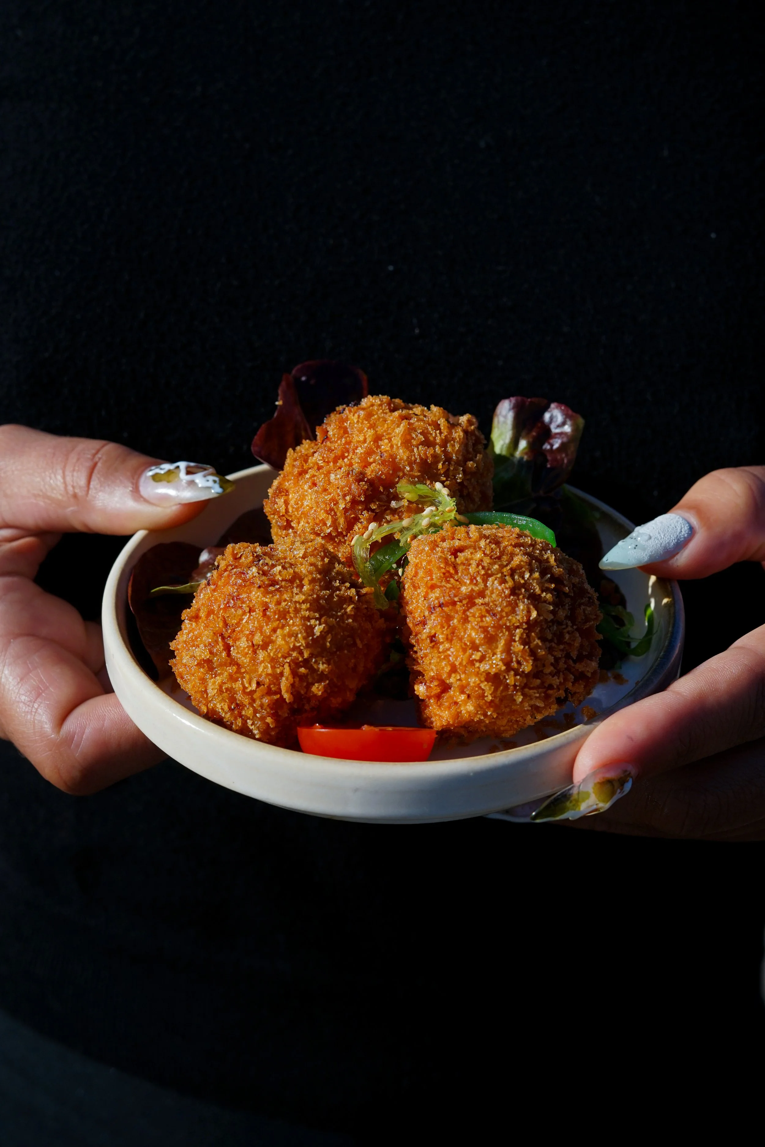 Gros plan de mains tenant un bol blanc contenant des croquettes frites, faites de viande ou poisson, accompagnées de légumes frais, avec un fond noir