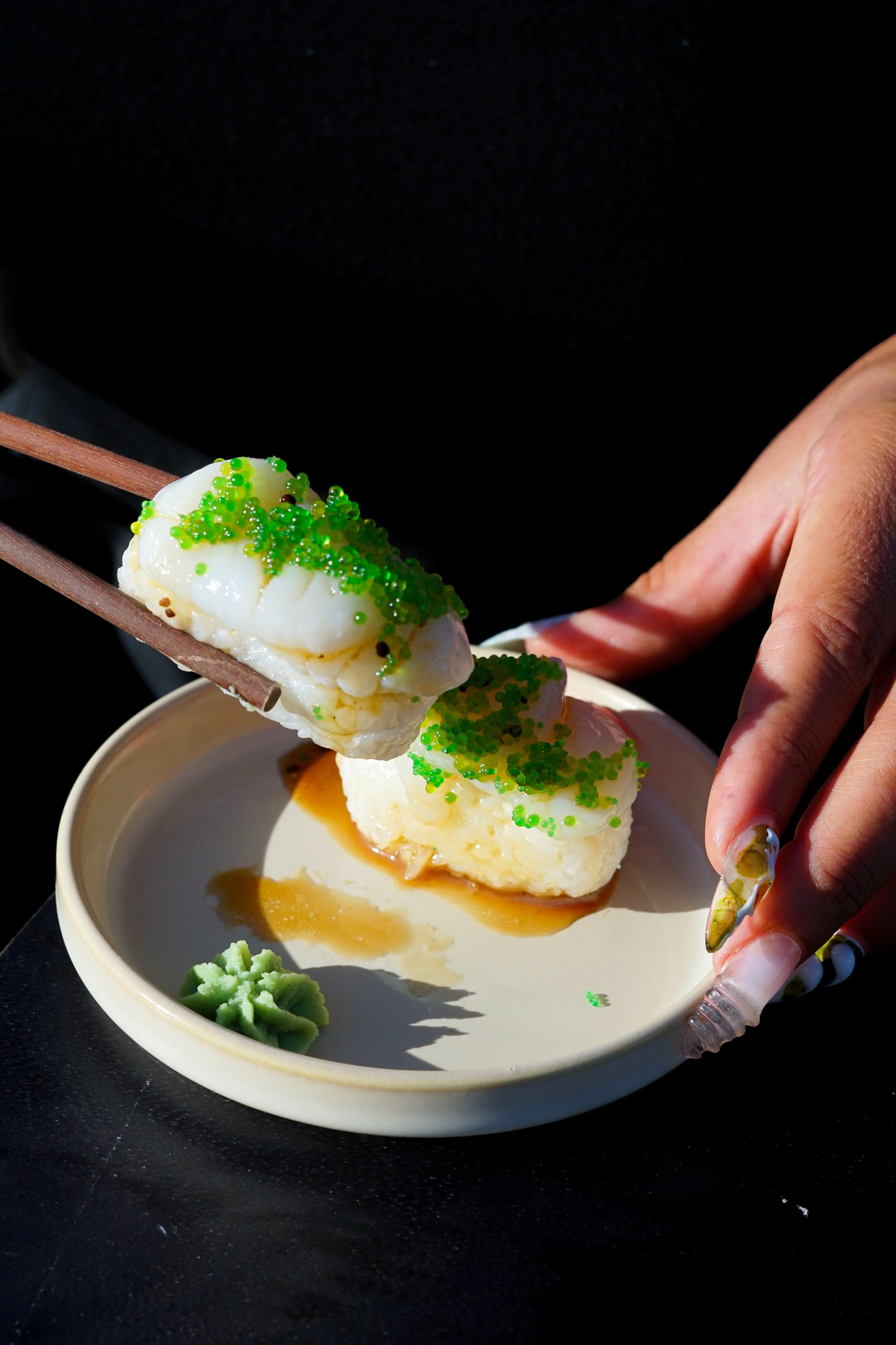 Une main tenant des baguettes avec une boule de glace à la vanille décorée de petites perles vertes. La glace est dans une petite assiette avec de la sauce caramel et un peu de wasabi à côté.