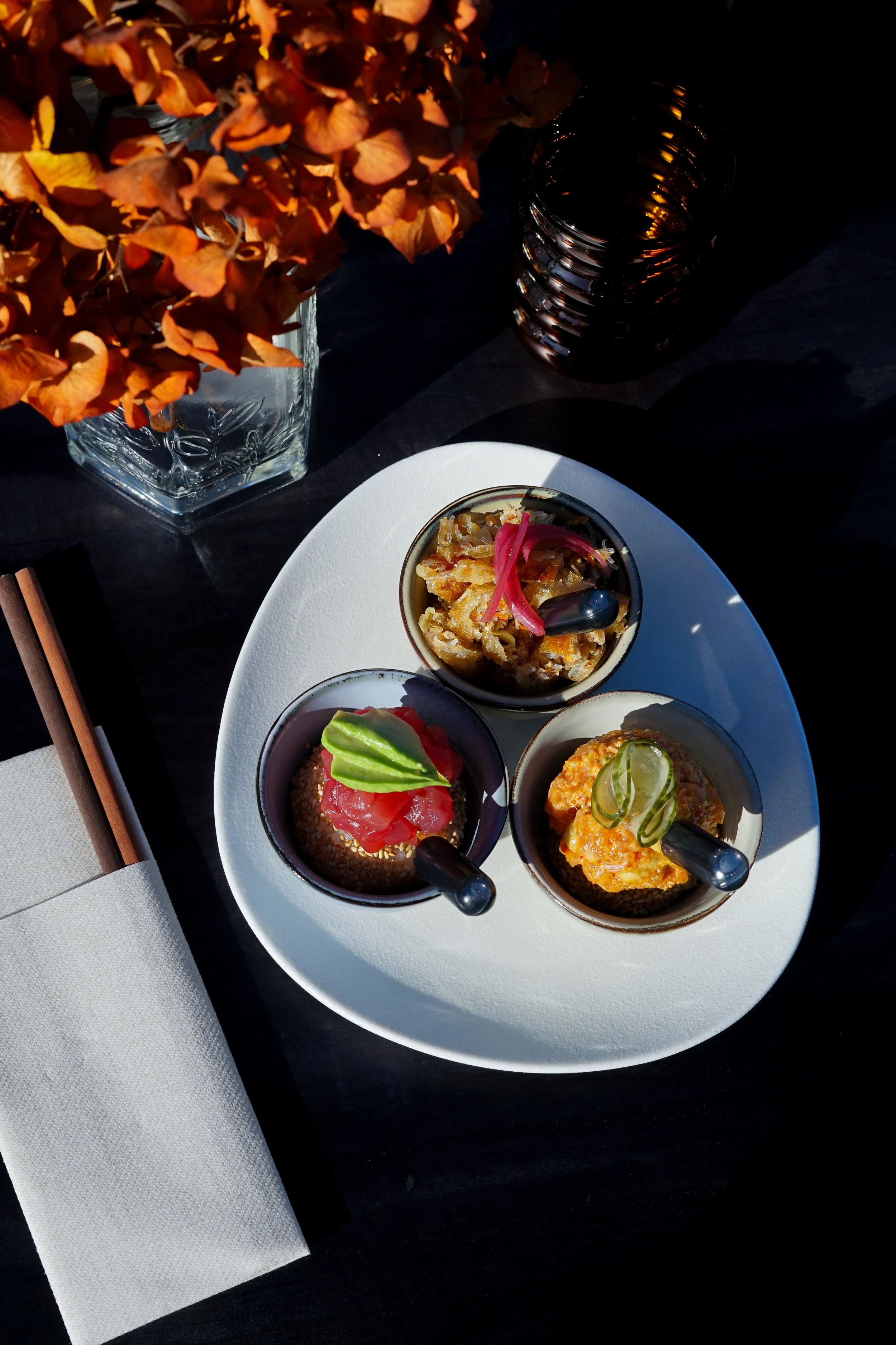 Assiette de petits plats japonais avec de la sauce, des légumes et de la viande, accompagnée d'une tasse de thé ou d'une boisson, et une bouteille en verre à côté, sur une table noire avec une nappe blanche et des baguettes.