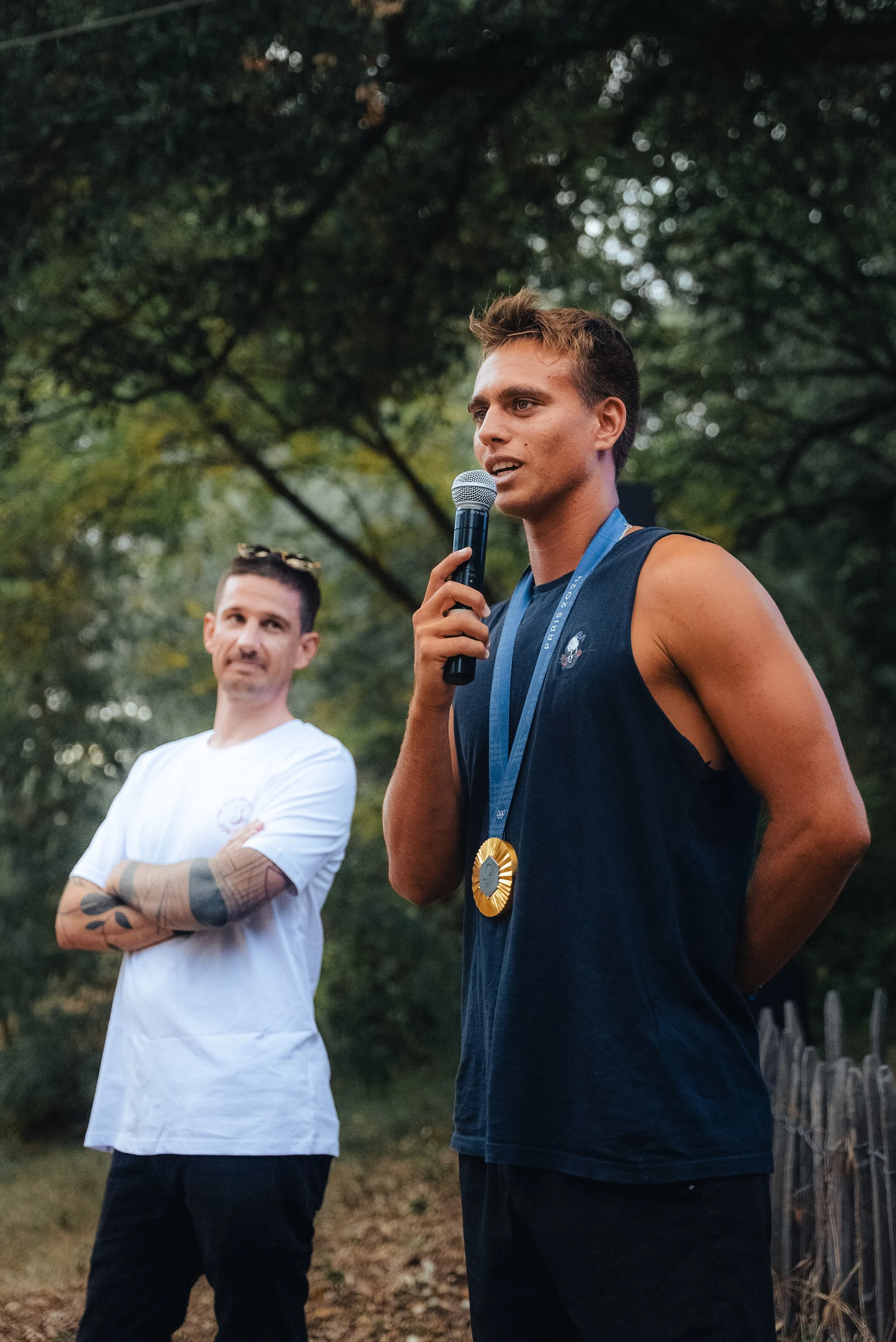 Deux hommes parlent lors d'une cérémonie en plein air, l'un portant une médaille. Arrière-plan d'arbres et une clôture en bois.