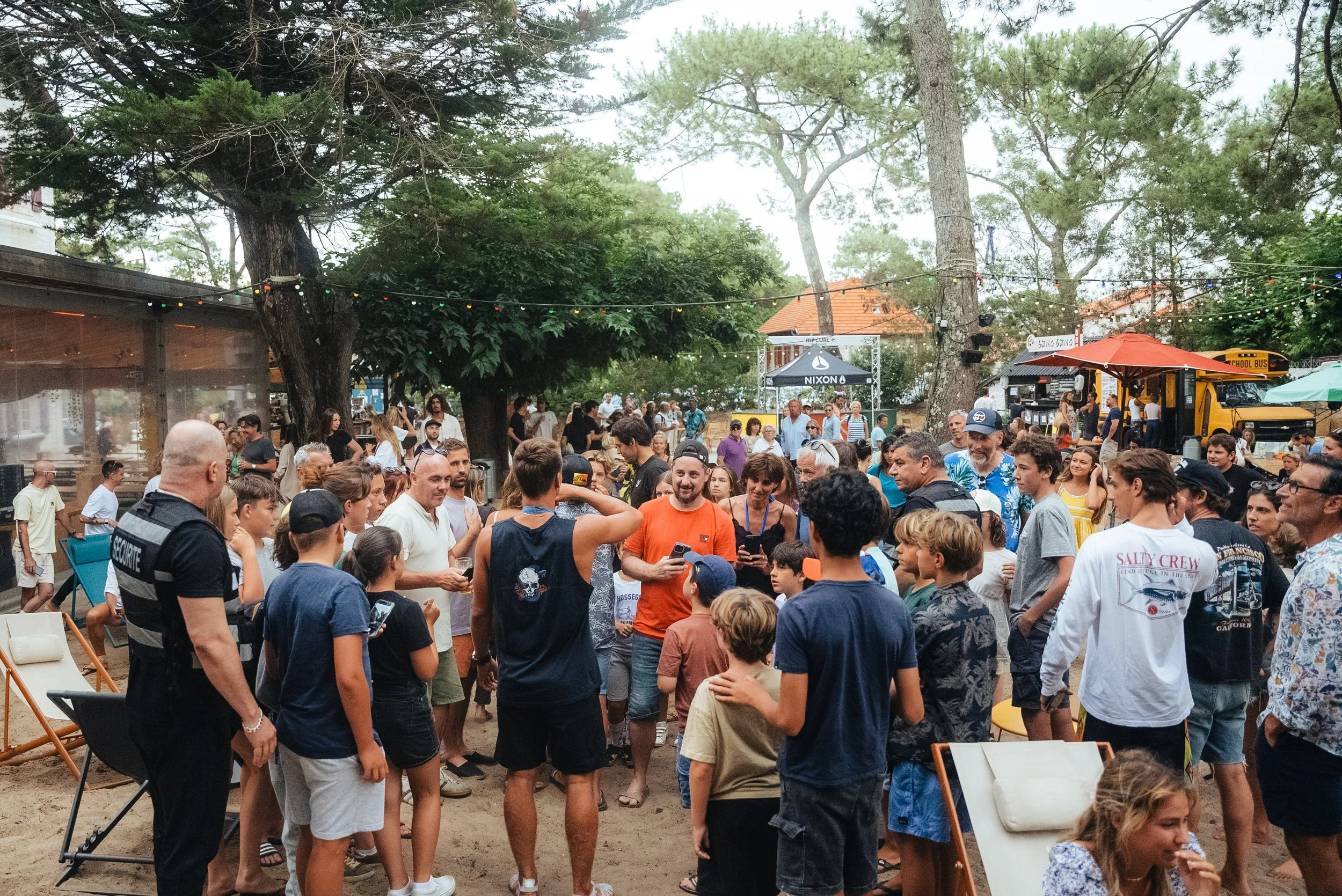 Une foule de personnes réunies en plein air lors d'un événement social ou festival, avec quelques stands, échafaudages et arbres en arrière-plan, sous un ciel nuageux.