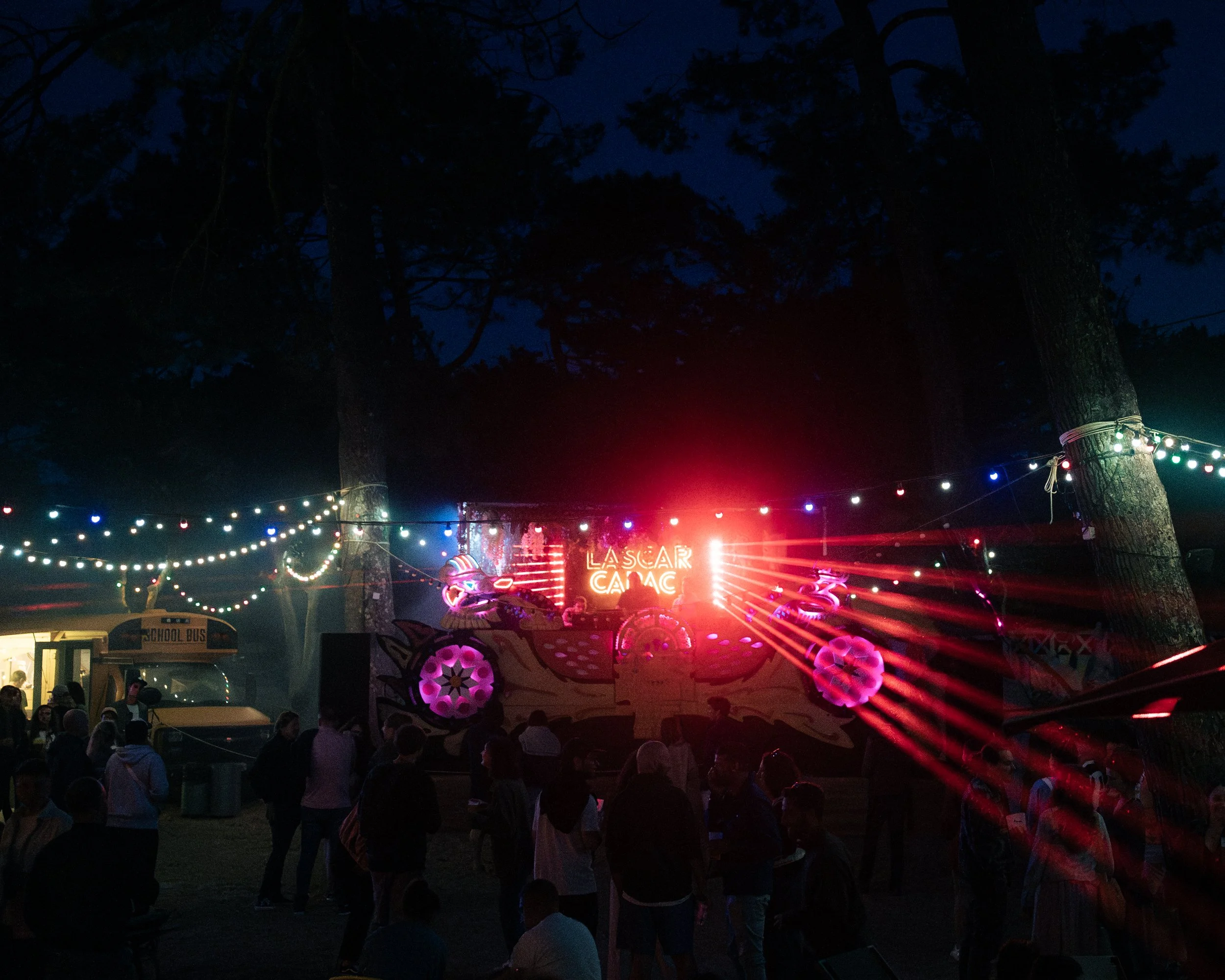 Une fête nocturne avec des lumières colorées accrochées entre deux grands arbres, une scène avec un panneau lumineux indiquant 'Lascar Cabac' et une foule de personnes rassemblées, avec un bus scolaire à l'arrière-plan.