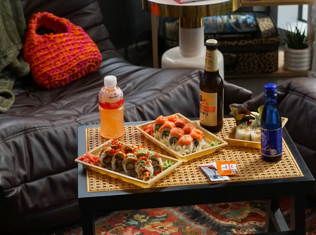 Une table avec des plateaux de sushis, une bouteille de bière, une boisson soda, un pack de soda, et une petite bouteille d'eau, dans un salon avec un canapé en cuir gris, un coussin en laine orange, une lampe et un tapis coloré.