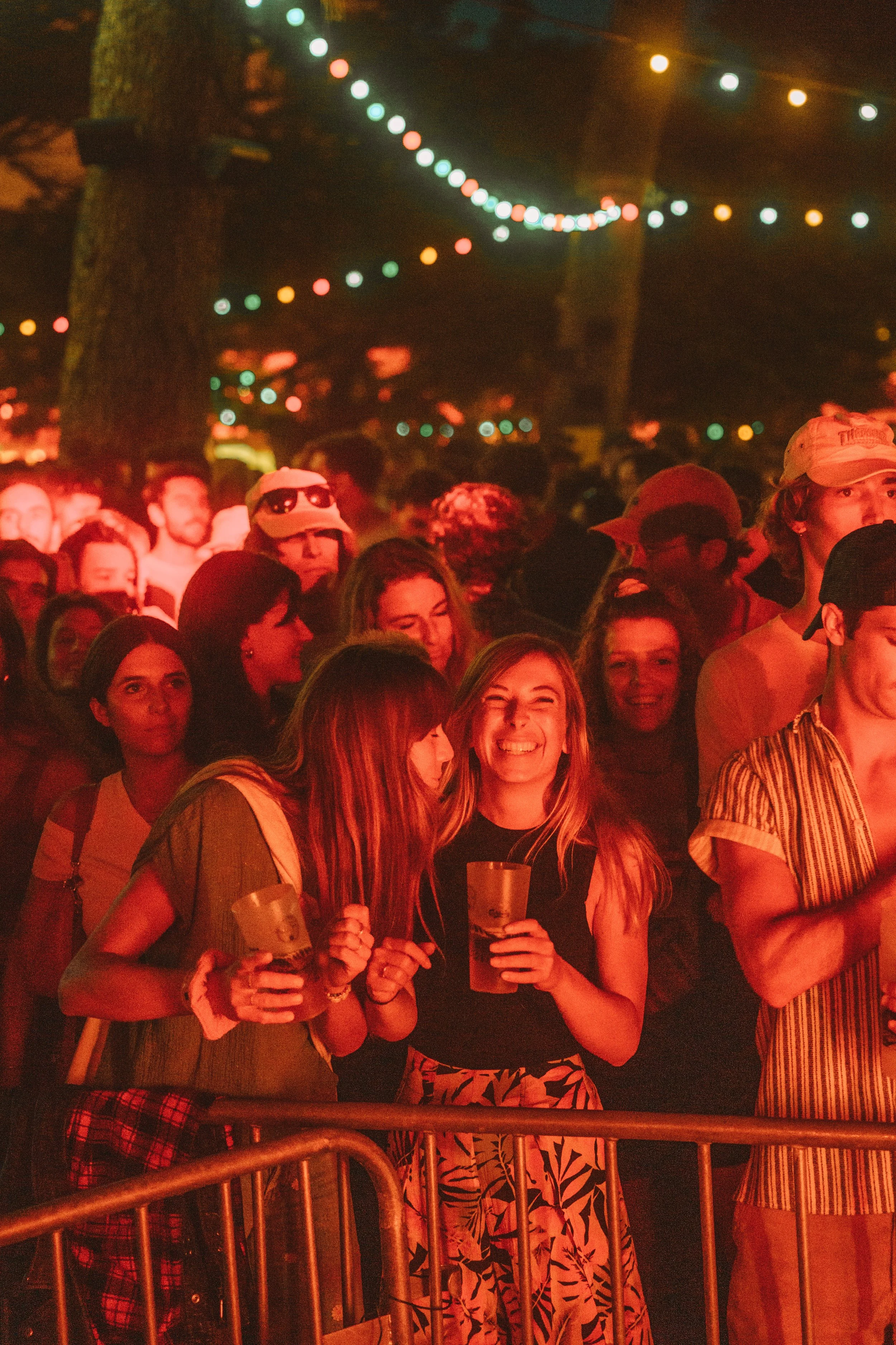Groupe de personnes assistant à un événement en plein air, éclairé par des guirlandes lumineuses, quelques personnes tiennent des verres en main, ambiance festive nocturne.