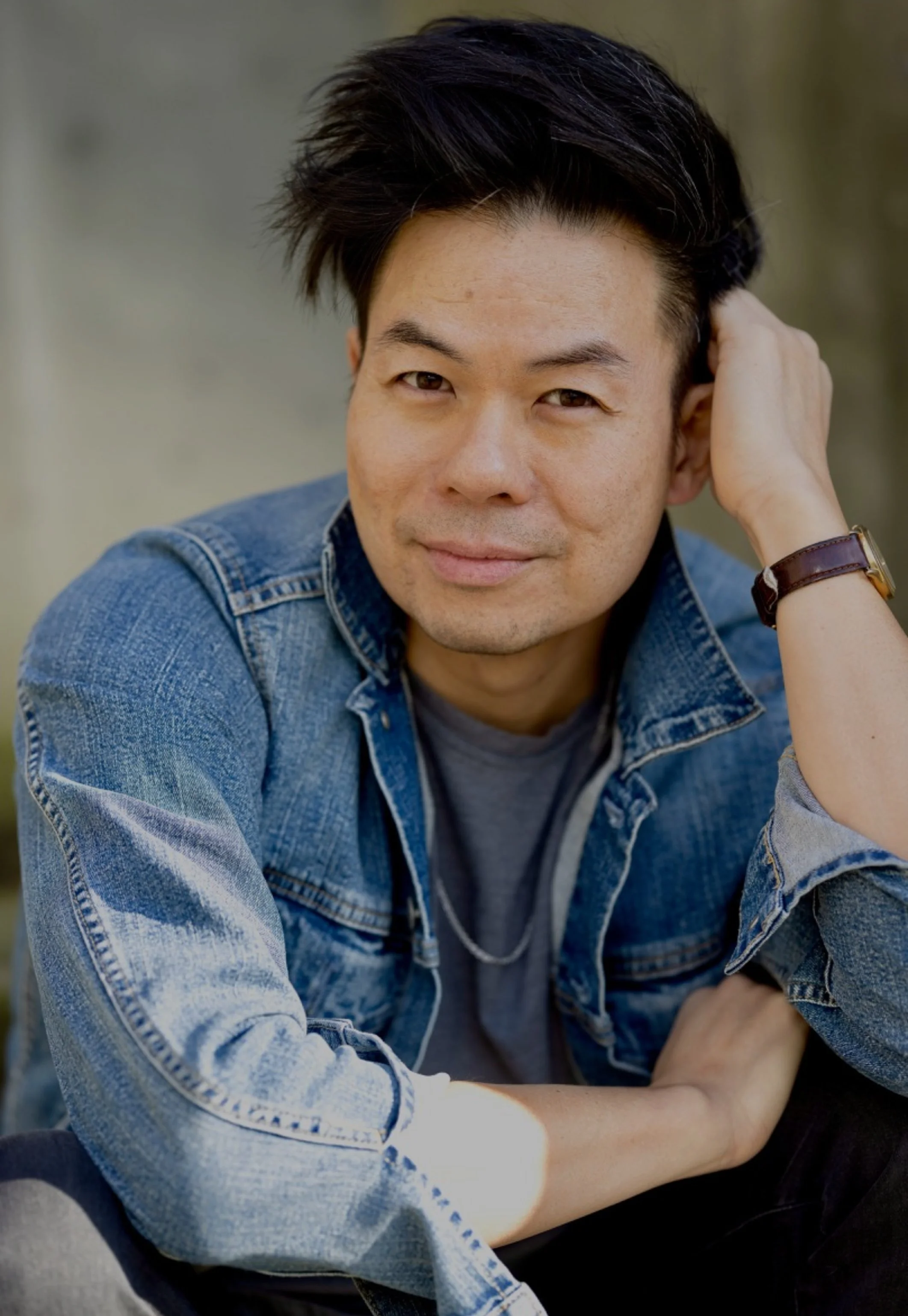 A young man with black hair, wearing a denim jacket and gray T-shirt, sitting outdoors with one hand behind his head, looking at the camera with a slight smile.