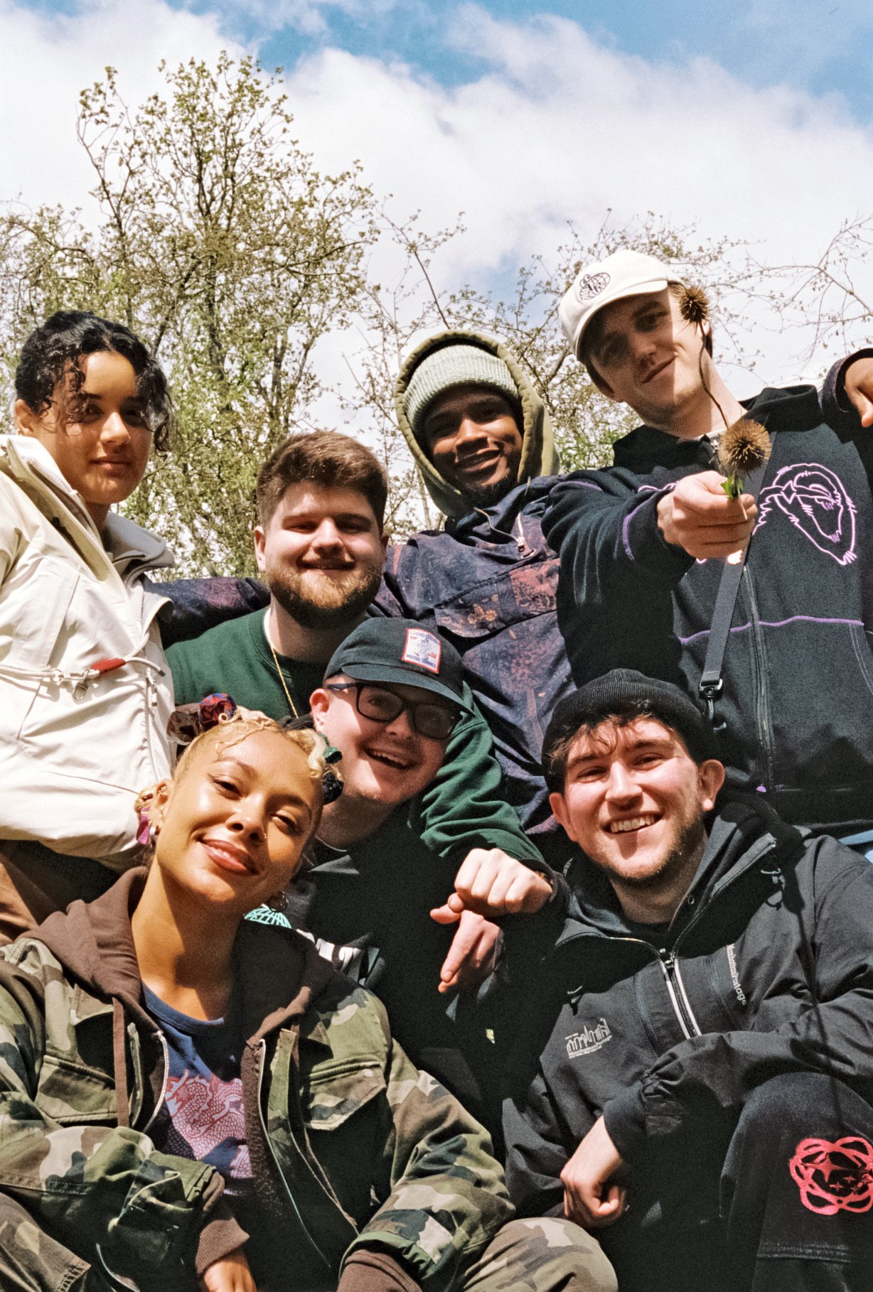 Musicians Iyamah, Kam-BU and Oma standing and crouching together for a photo, smiling outdoors on a sunny day, some wearing jackets and hoodies, with trees and a partly cloudy sky in the background.