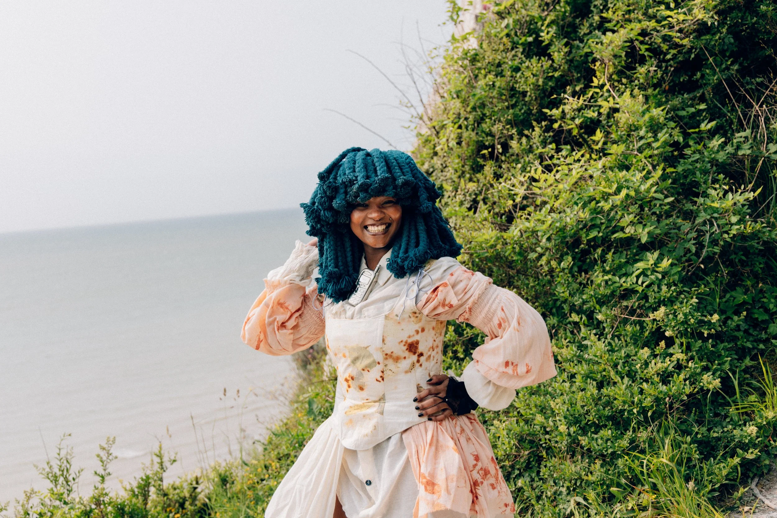 Musician Moonchild Sanelly posing in front of greenery with the sea in the background, with her blue braided hair, wearing a floral patterned dress with long, flowy sleeves, smiling with one hand on her waist and the other lifted up to hear head.