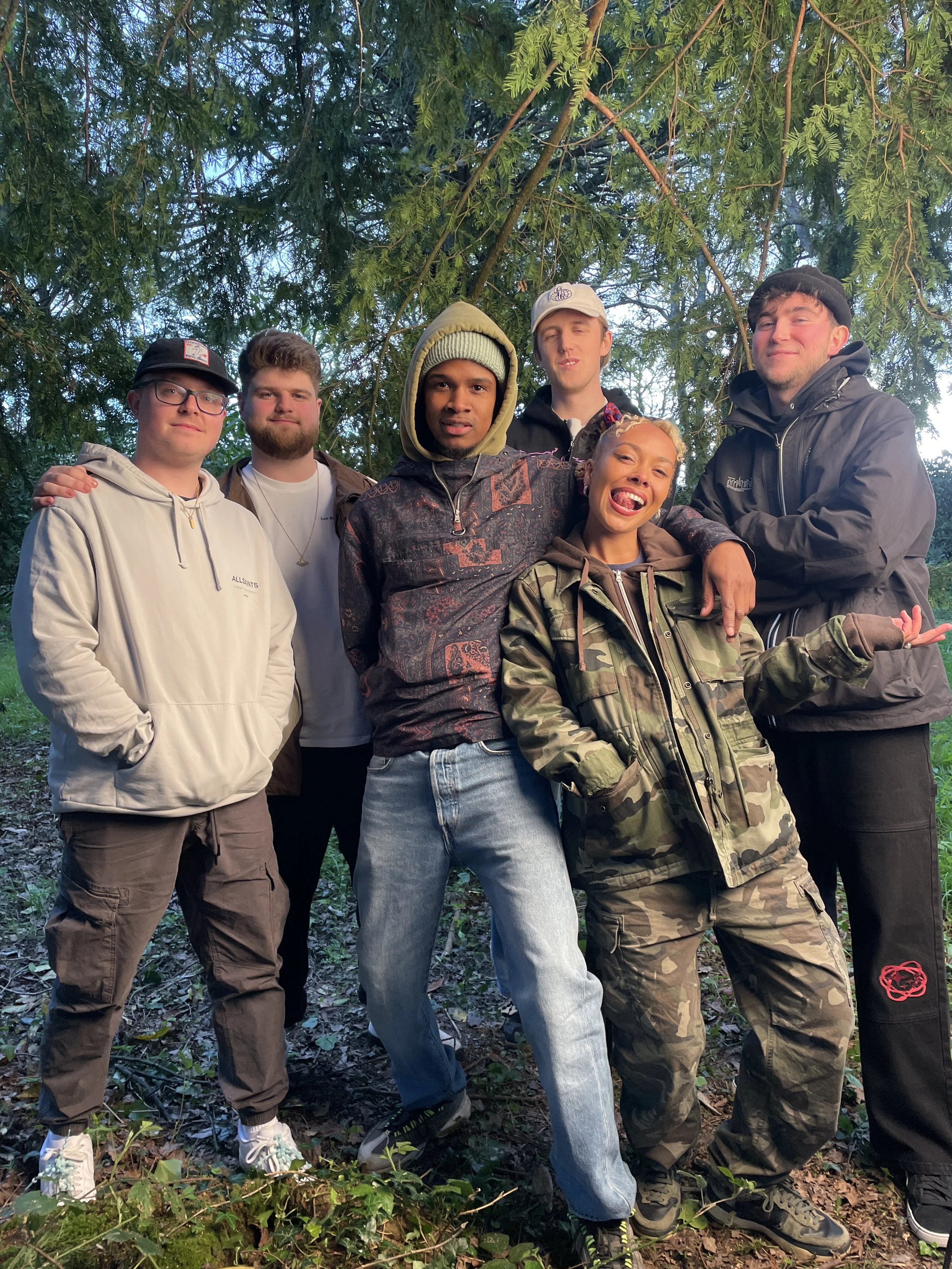 Musicians Iyamah, Kam-BU and Oma standing together for a photo, smiling outdoors on a sunny day, some wearing jackets and hoodies, with trees in the background.