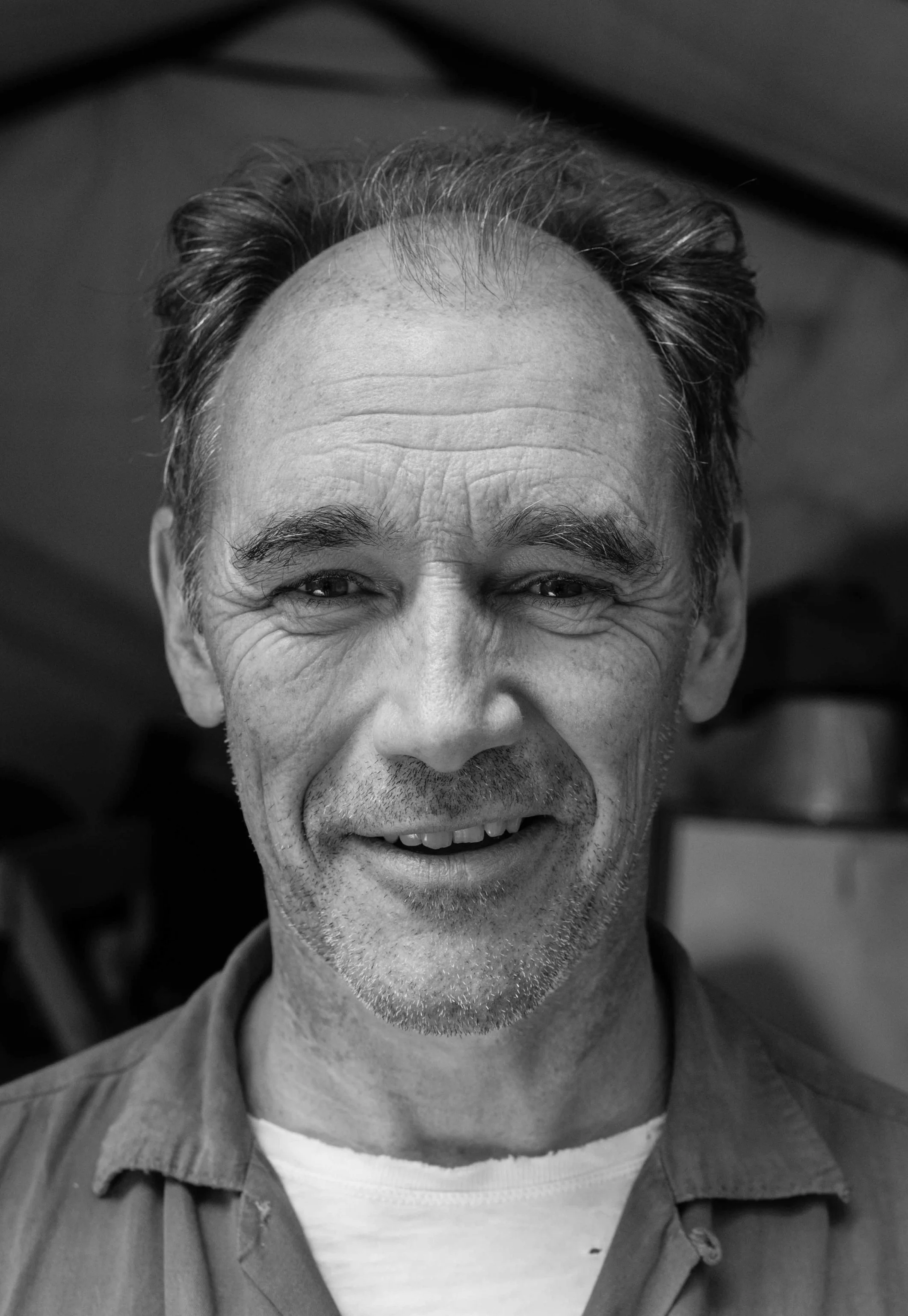 Black and white headshot of Mark Rylance smiling with short, wavy hair and a beard, wearing a collared shirt.